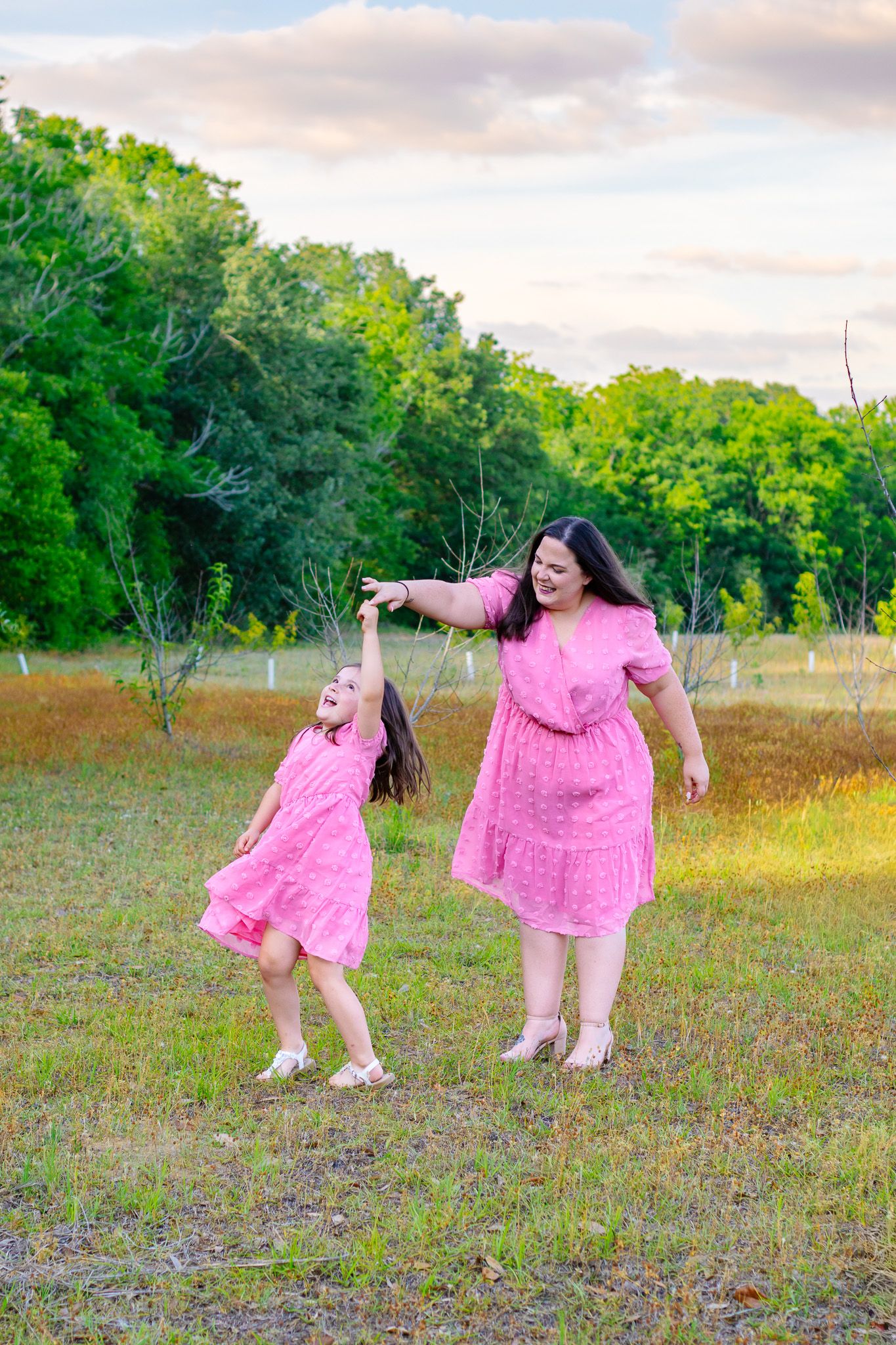 Mother and daughter in matching pink dresses dancing joyfully in a green field at sunset.