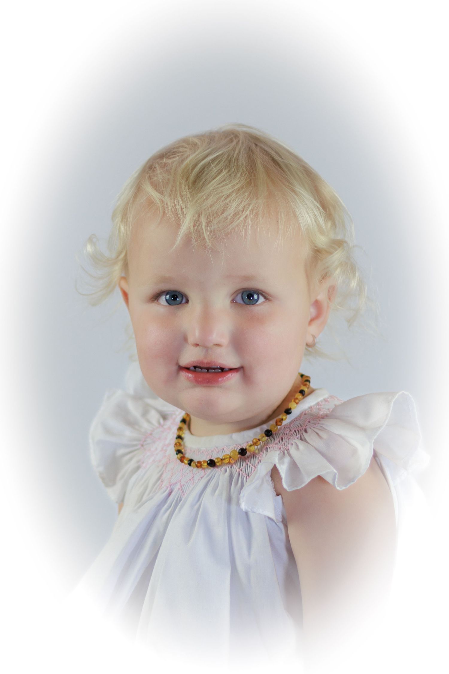 Adorable blonde toddler girl with blue eyes wearing a white dress and amber bead necklace, smiling in a portrait photo.
