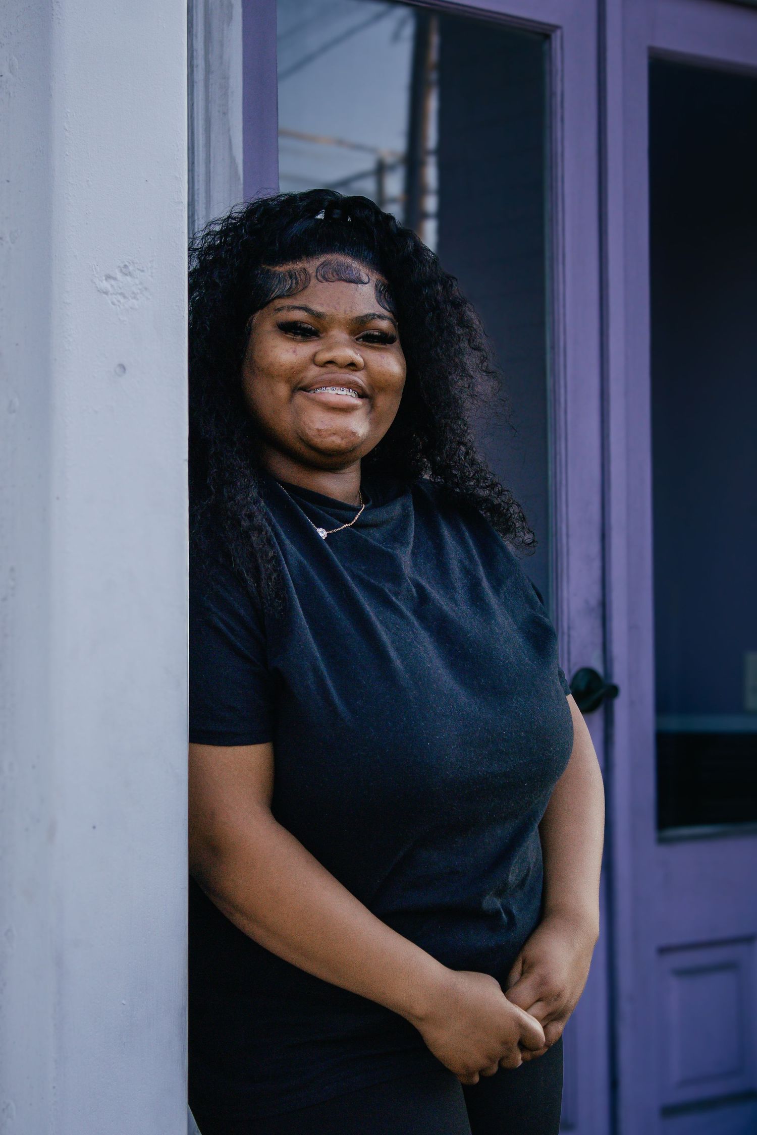 Young Black woman with curly hair smiling in a black t-shirt, leaning against a purple door frame.