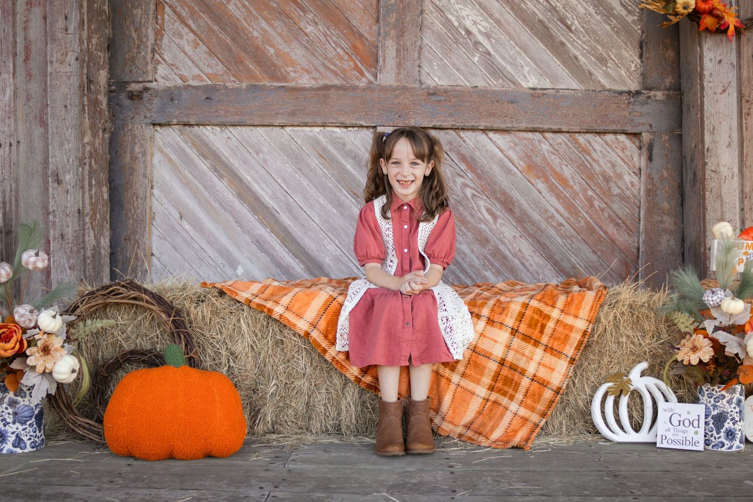 Young girl in red dress sitting on hay bale with pumpkins in autumn barn setting.