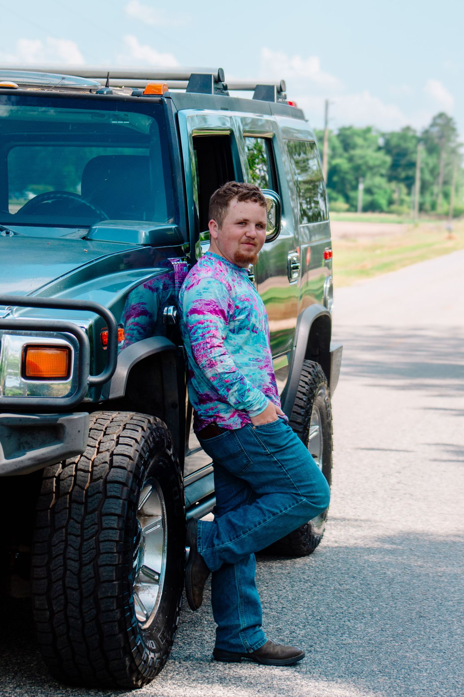Man in colorful tie-dye shirt and jeans leaning against a green Hummer H2 on a rural road.