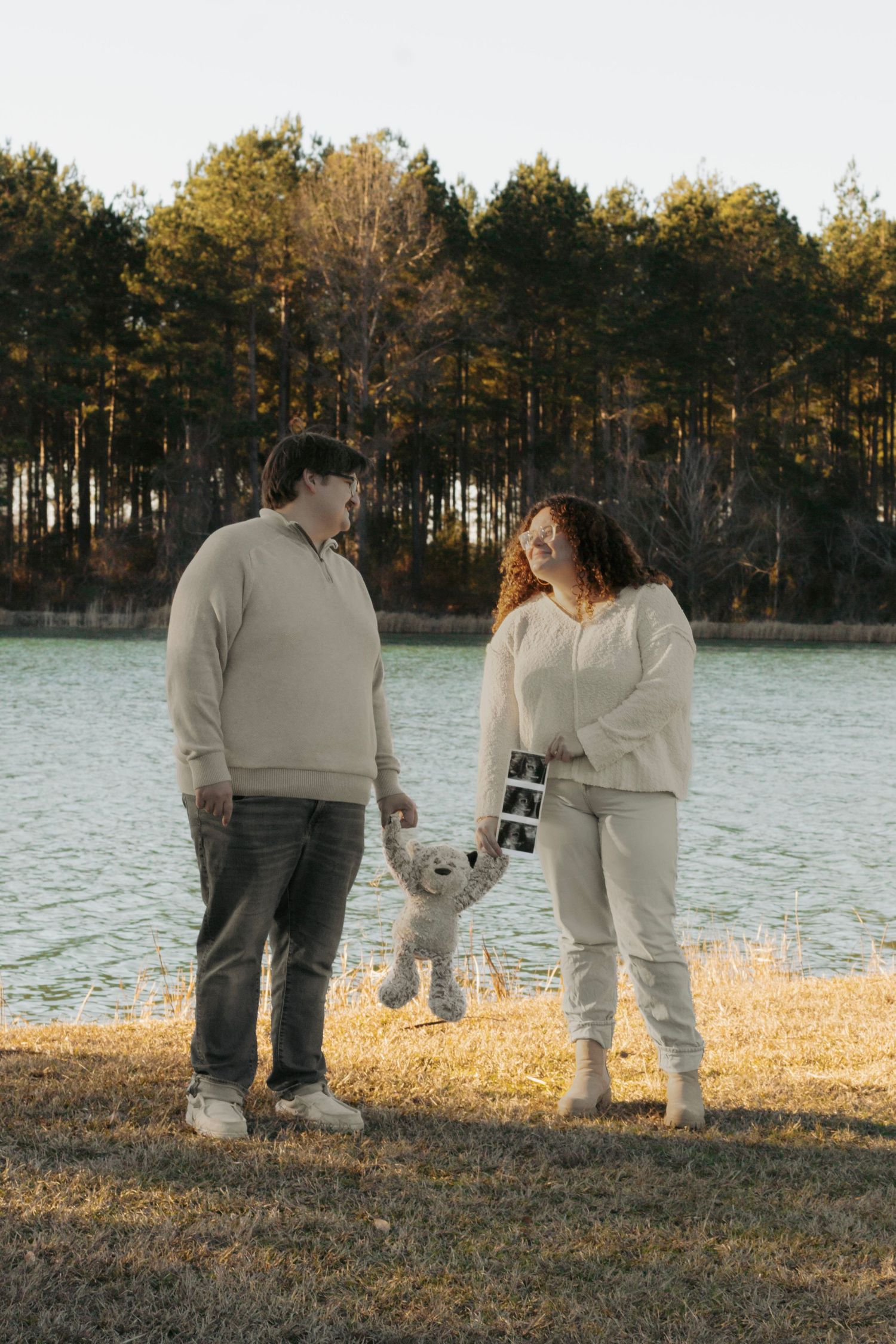 Couple holding teddy bear and ultrasound photos by a lake at sunset.