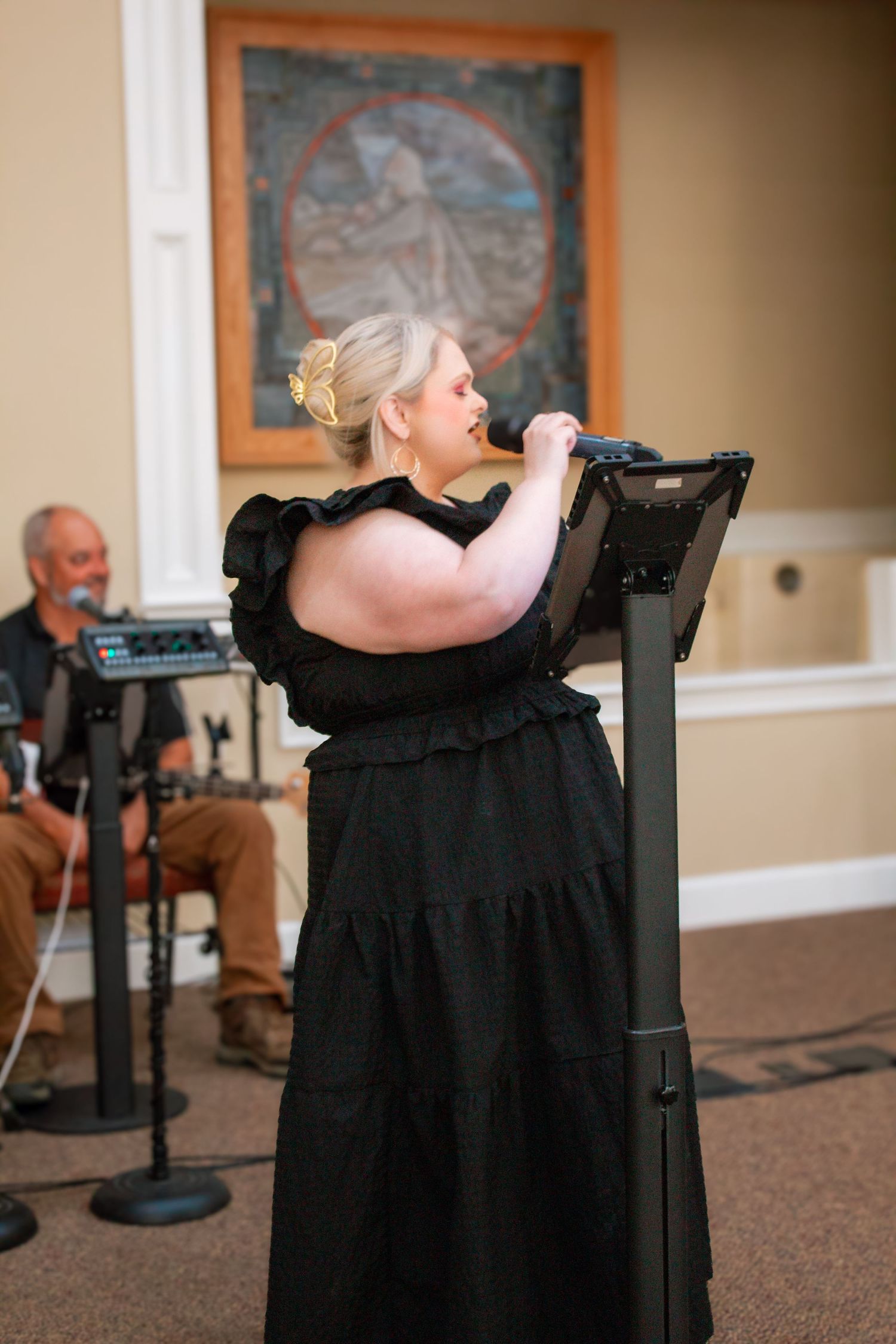 A blonde woman in a black ruffled dress performs vocals at a church event with a musician behind her.