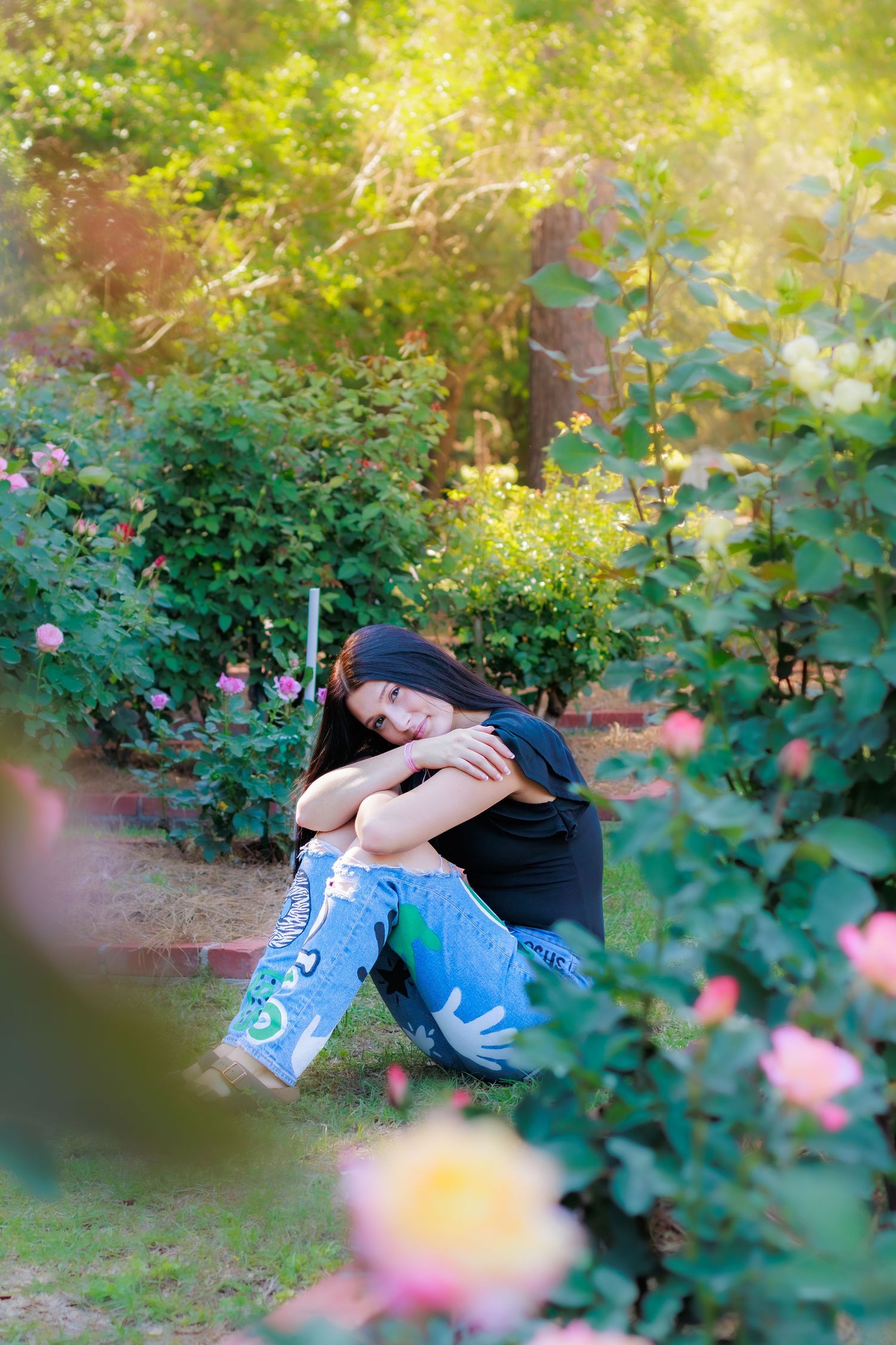Young woman with dark hair sitting in a garden surrounded by colorful roses wearing painted jeans.
