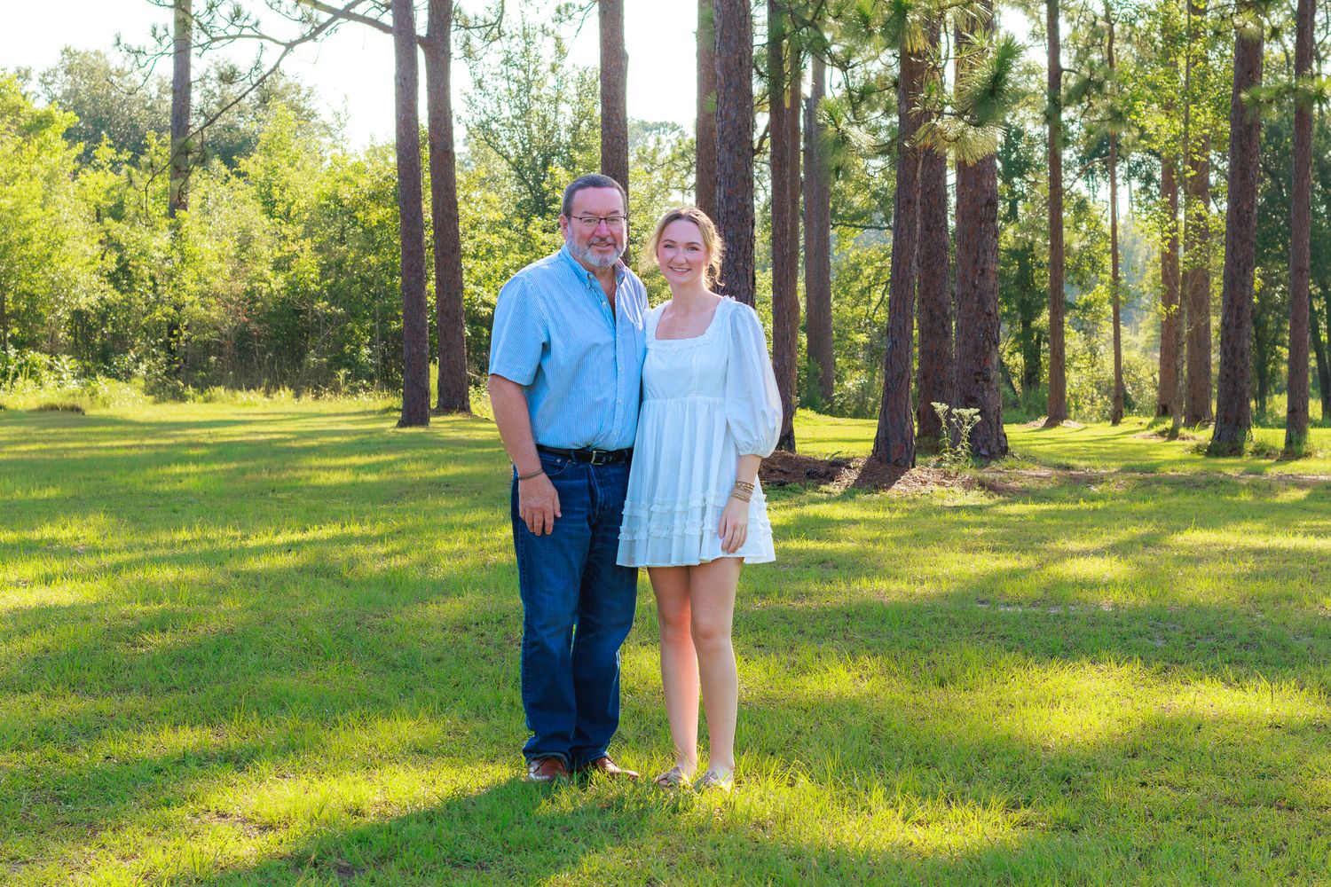 A couple smiles together on a green lawn surrounded by tall pine trees in a sunny outdoor setting.