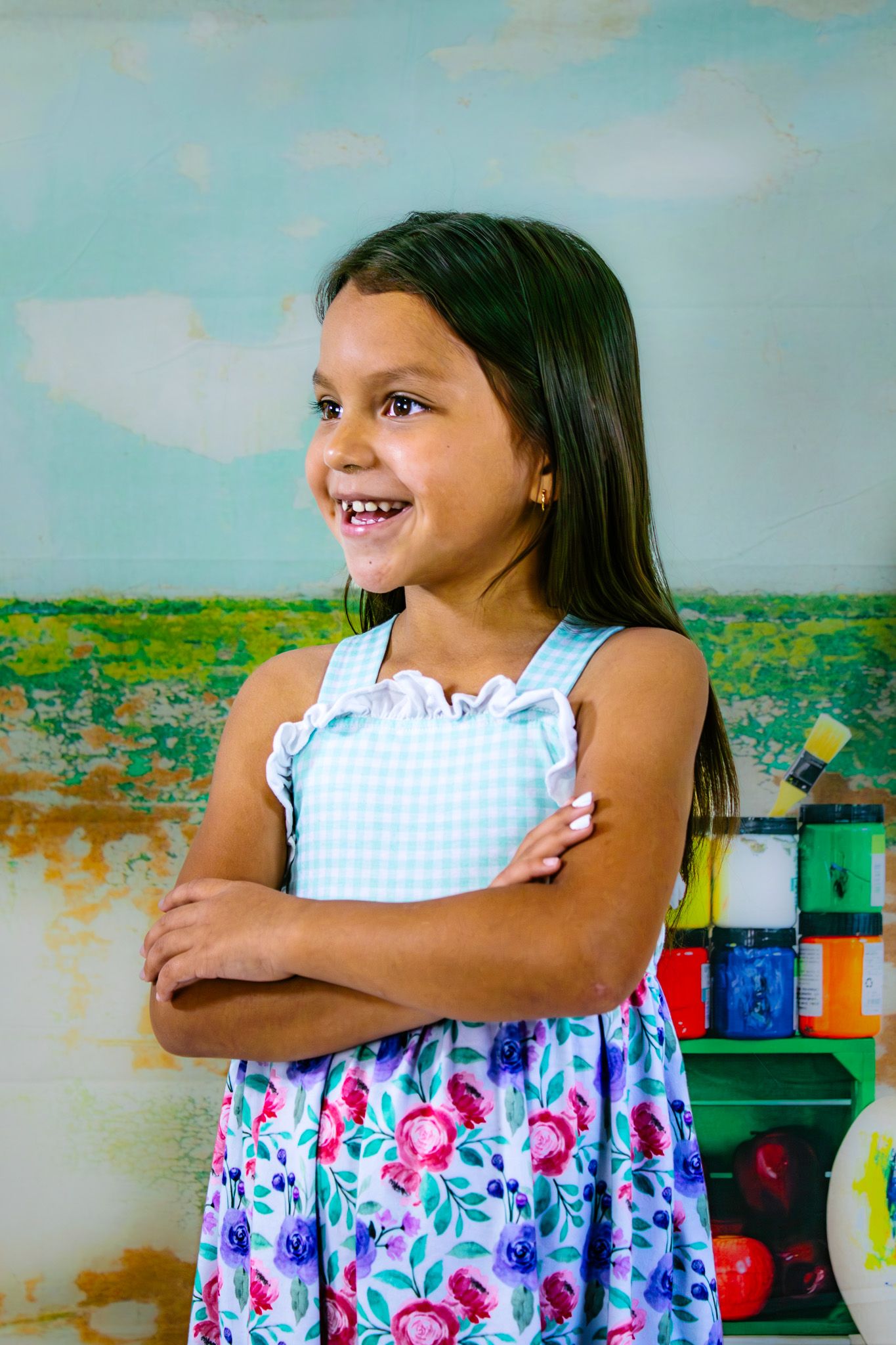 A smiling young girl in a floral dress stands with arms crossed in a colorful classroom setting.