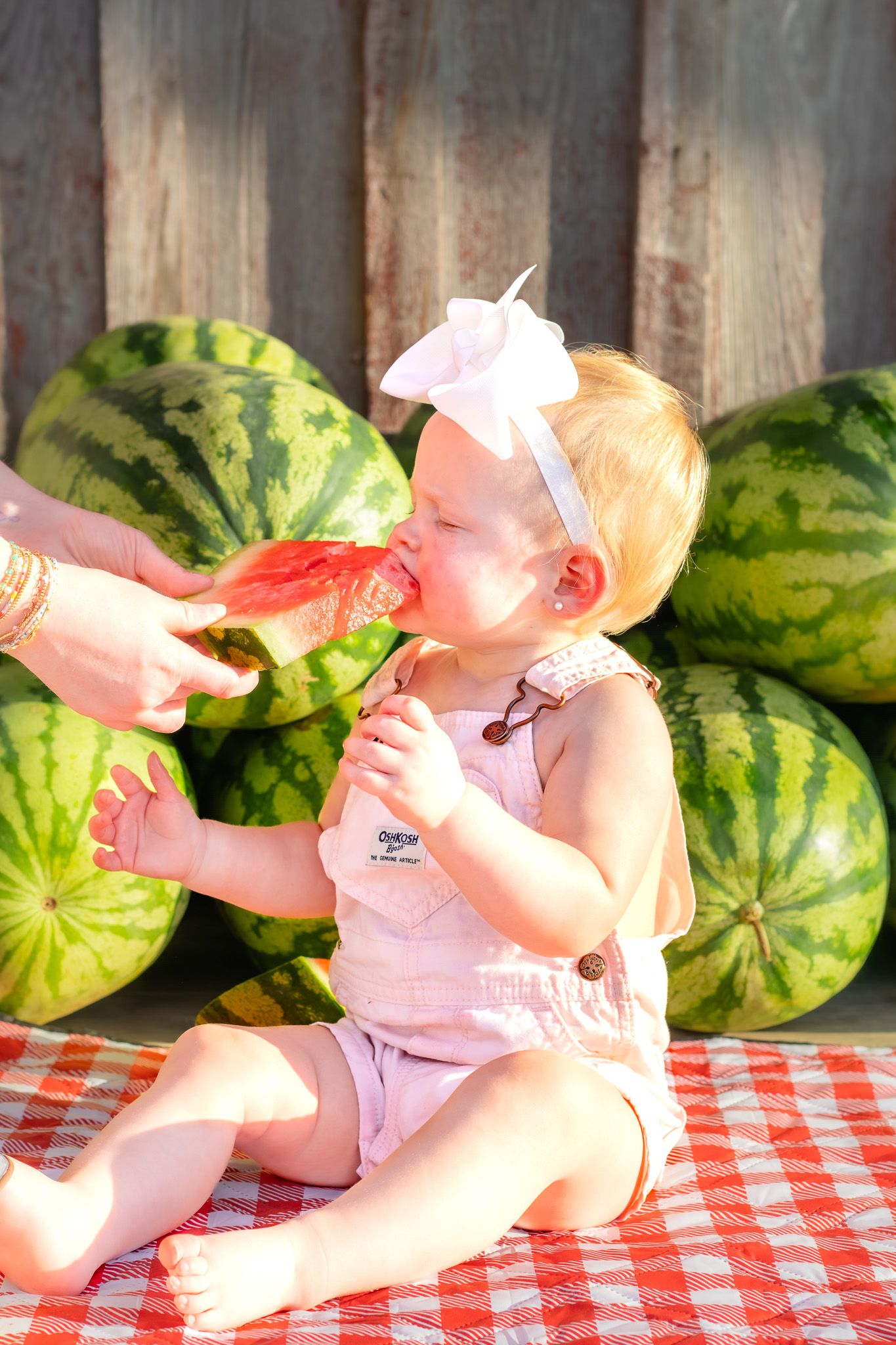 Adorable baby girl eating fresh watermelon slice outdoors, sitting on checkered blanket surrounded by whole watermelons.