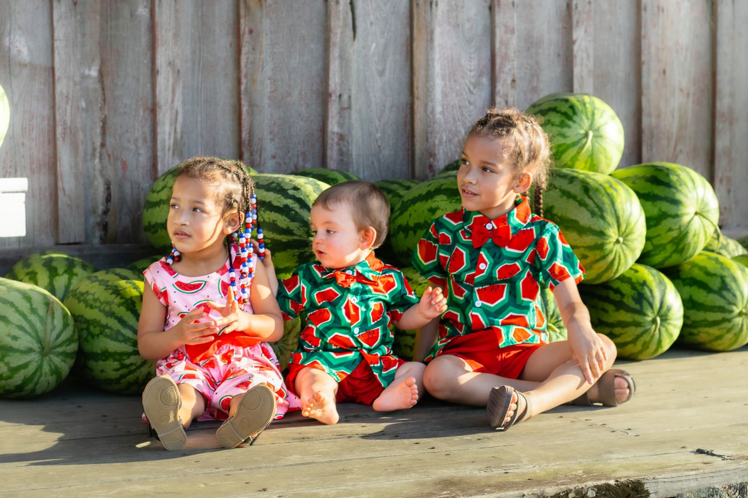 Three young children in watermelon-print outfits sit together on wooden steps surrounded by large watermelons.