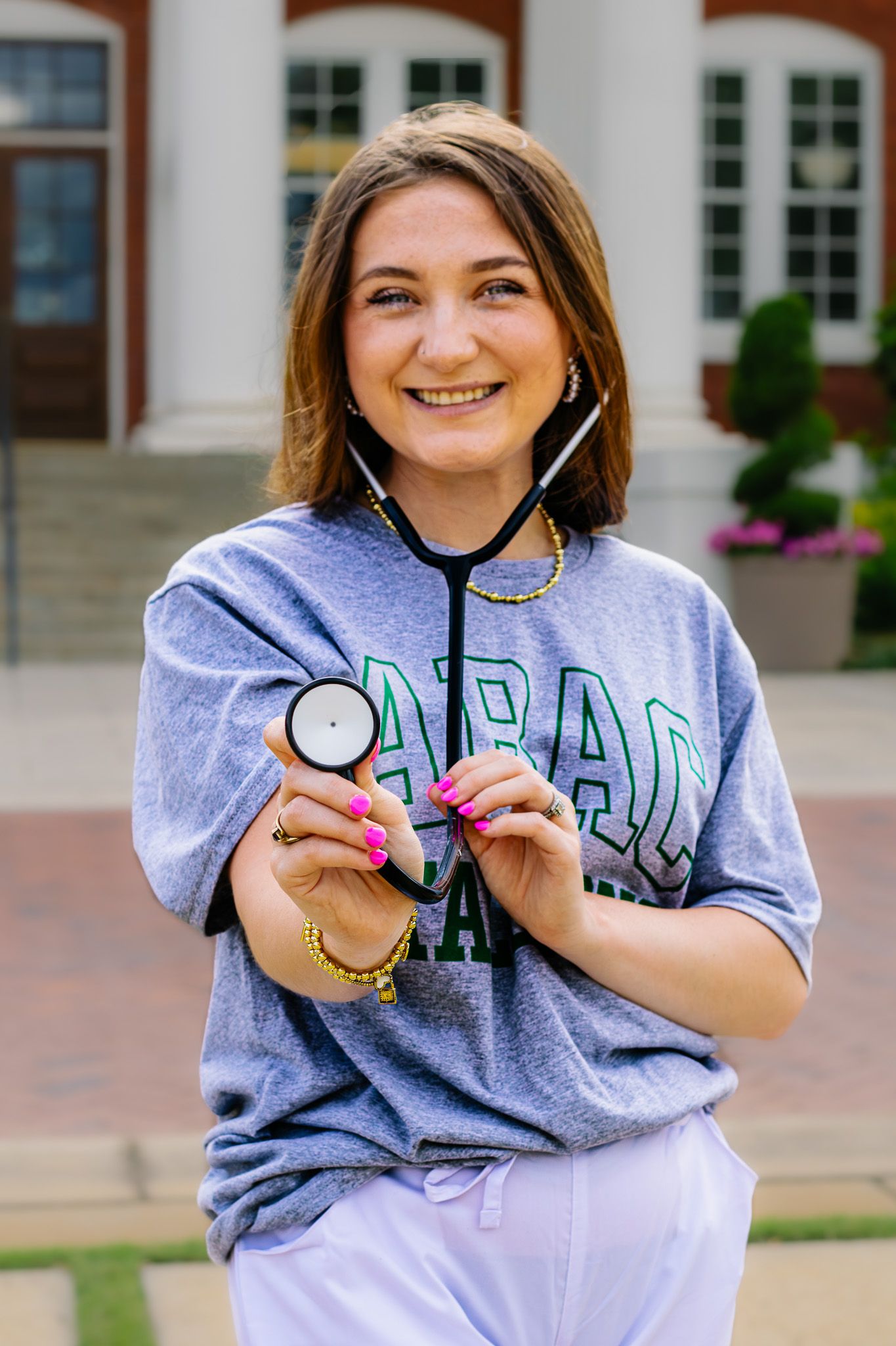 Smiling young woman in college shirt holds stethoscope outside brick university building.