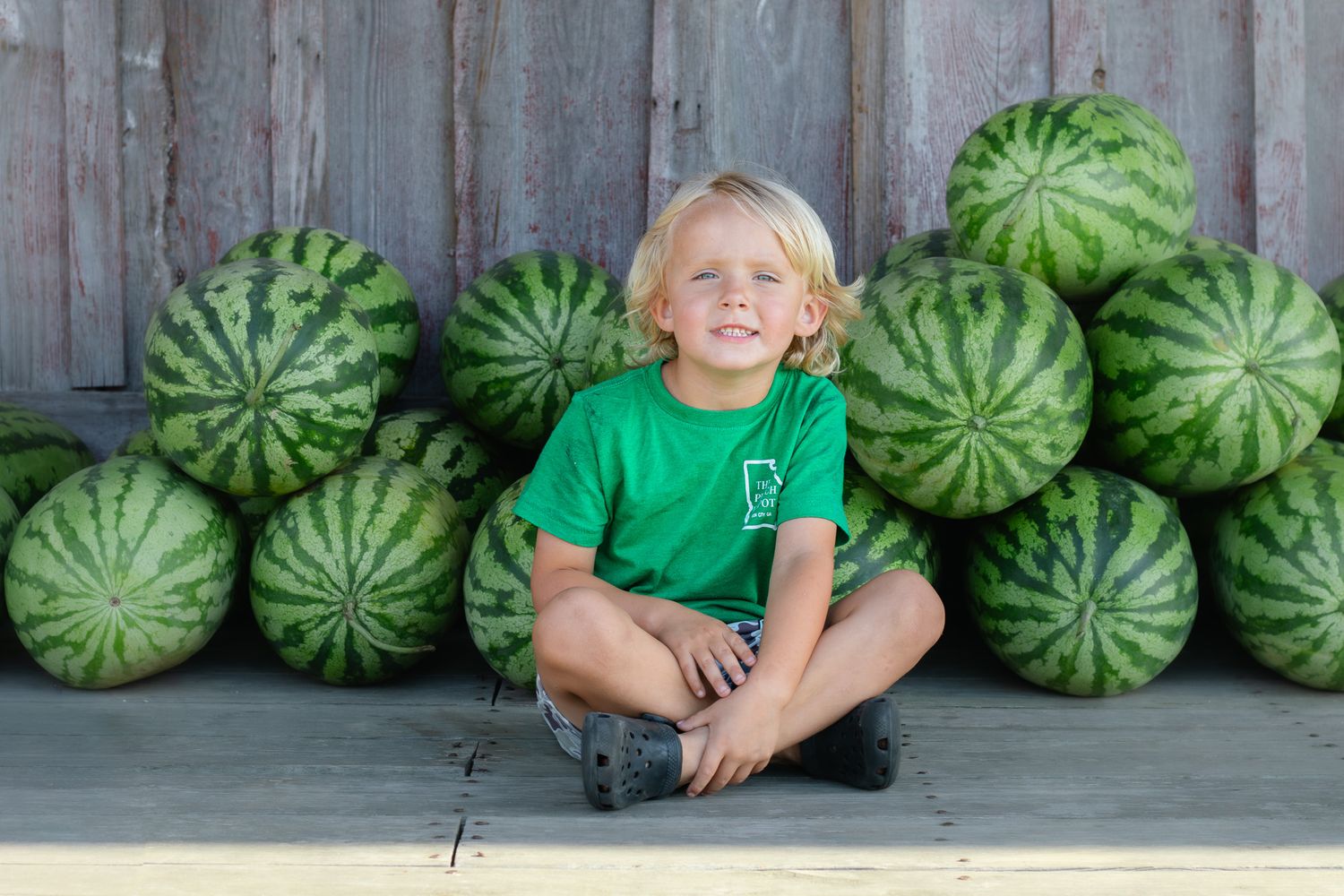 Smiling blonde boy in green shirt sitting cross-legged in front of a large pile of watermelons on a wooden surface.