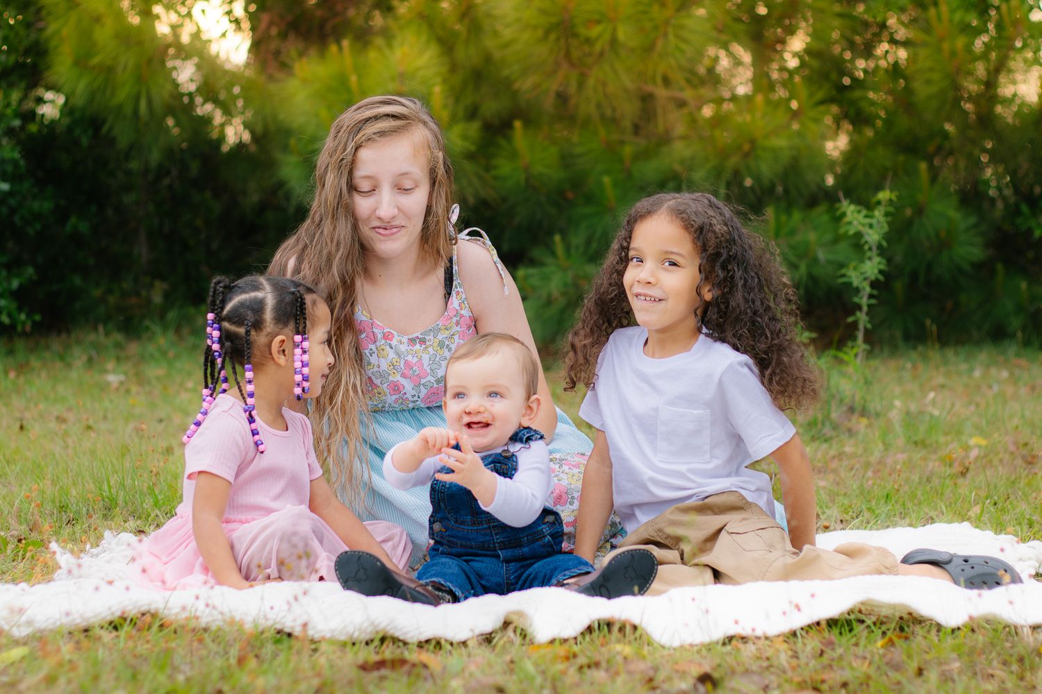 Teen girl sitting on blanket in grass with three young children on sunny day outdoors.