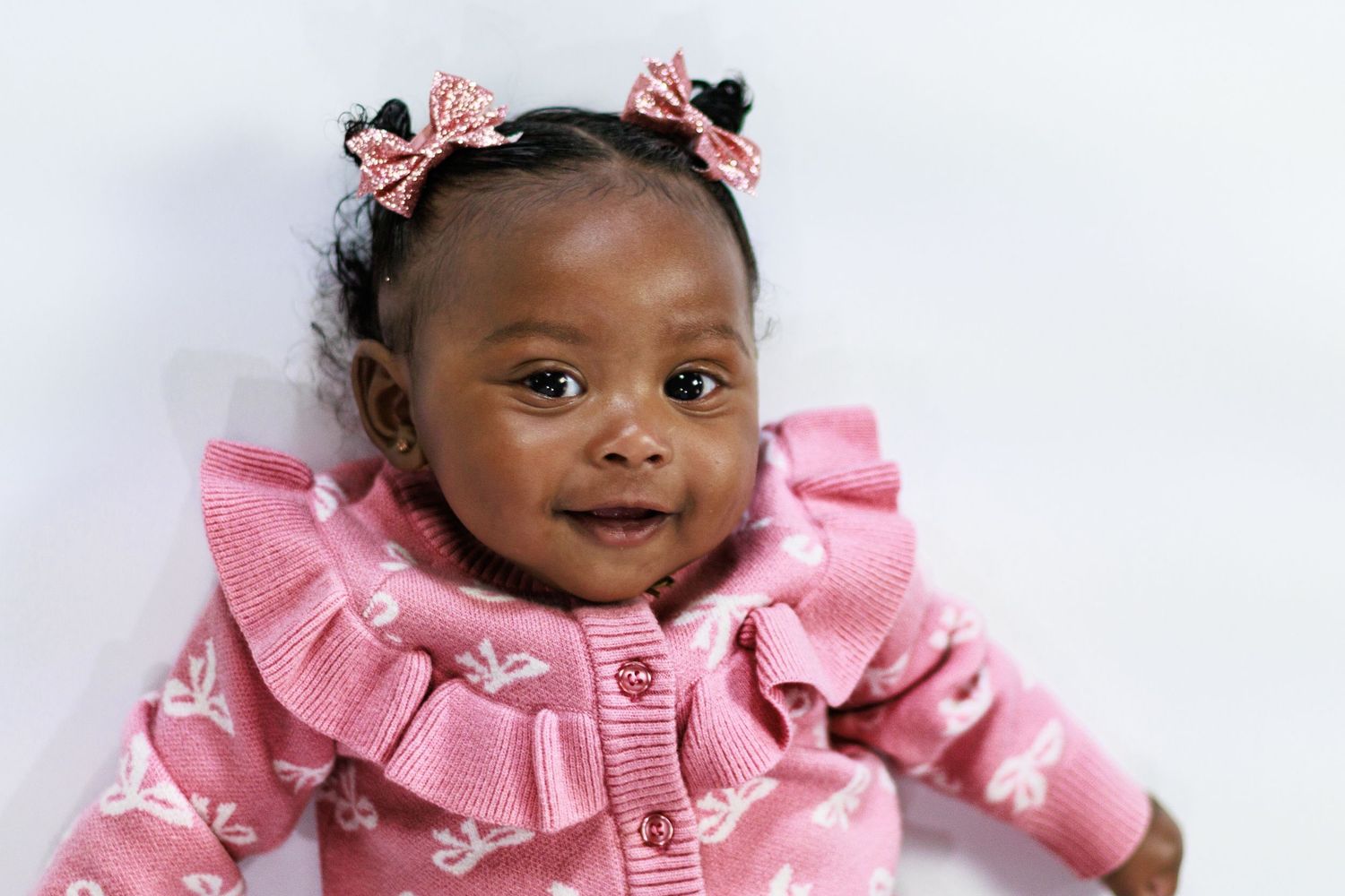 Smiling baby girl in pink ruffled outfit with floral bows in her hair, lying on a white background.