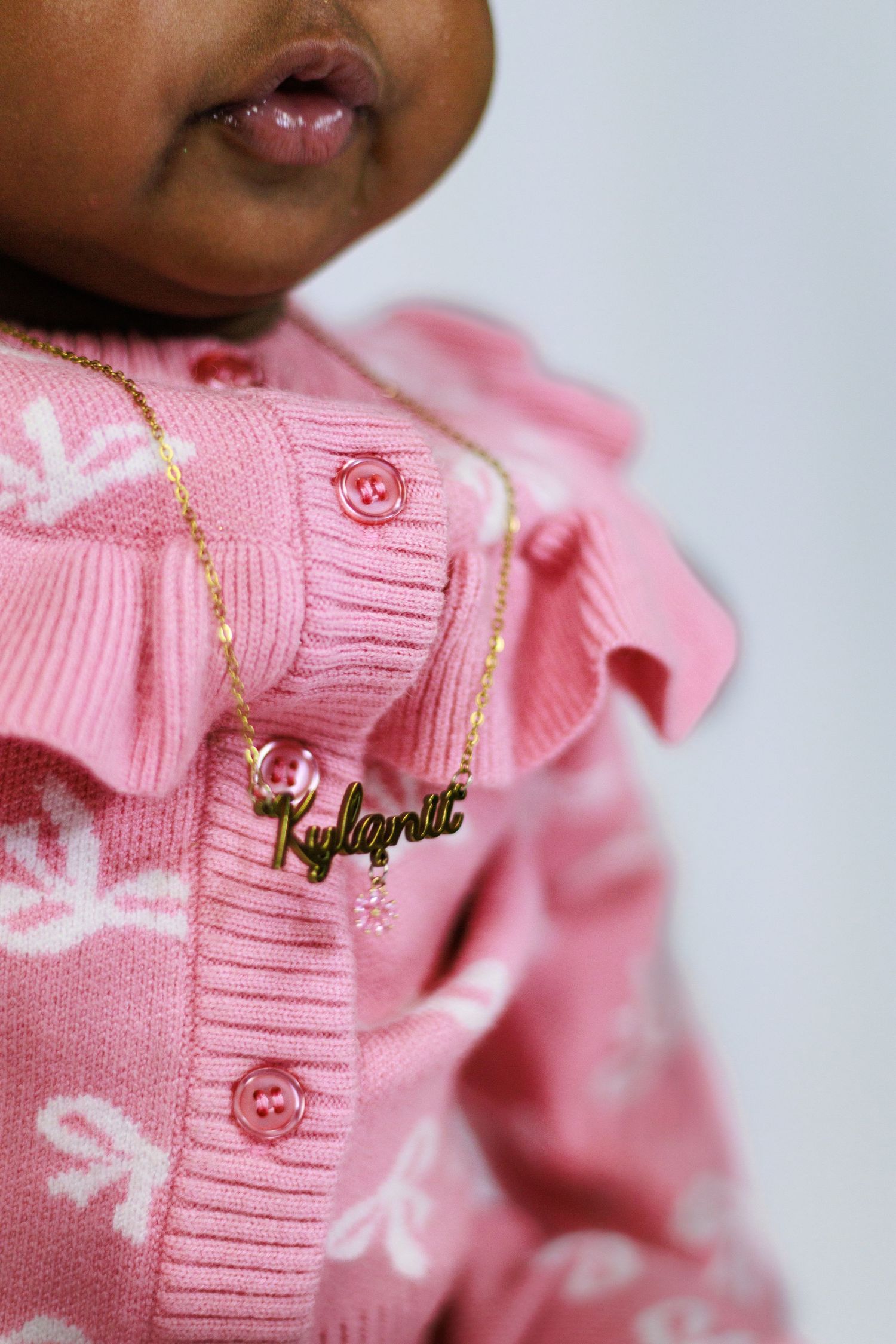 Baby wearing pink knit cardigan with ruffles and gold nameplate necklace reading Kylarni.