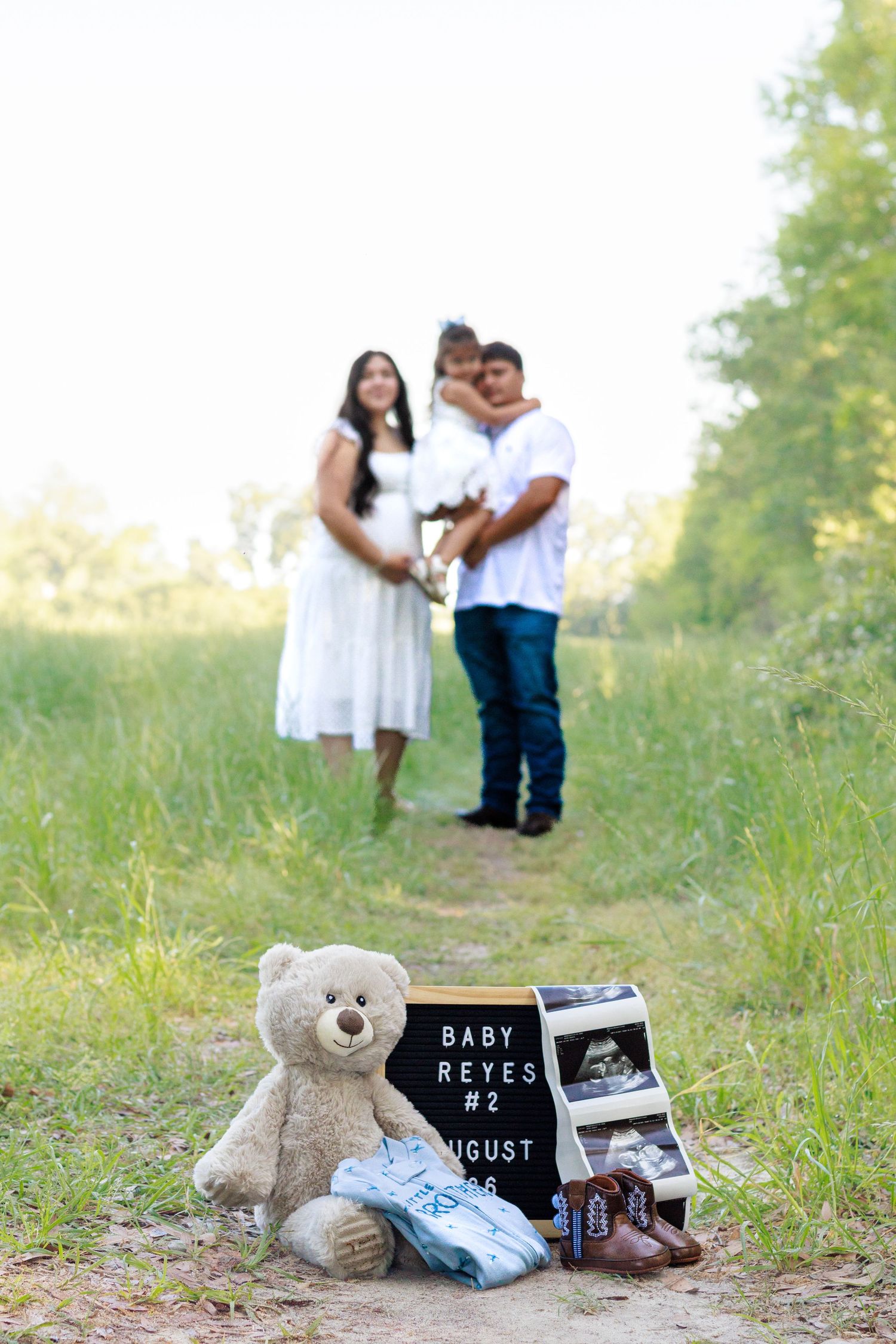 Family pregnancy announcement for Baby Reyes #2 with teddy bear, sonogram, and tiny shoes on a nature path.