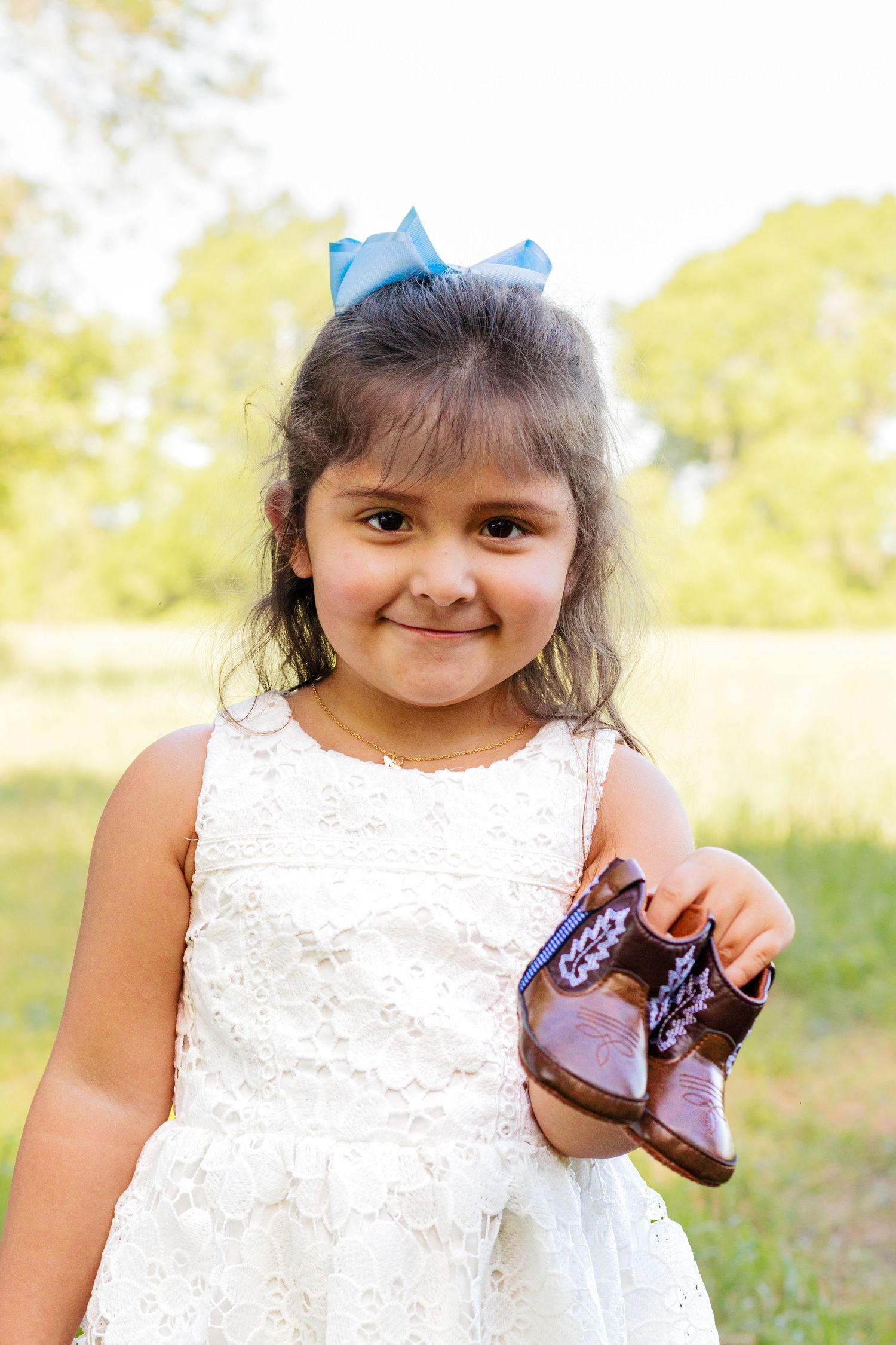 Smiling little girl in white dress holds baby shoes with blue bow in hair outdoors.