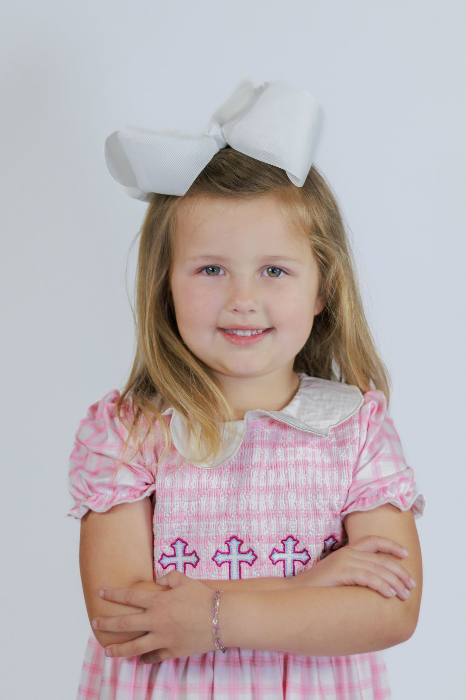 Young girl in pink plaid smocked dress with cross embroidery and white hair bow, arms crossed, smiling.
