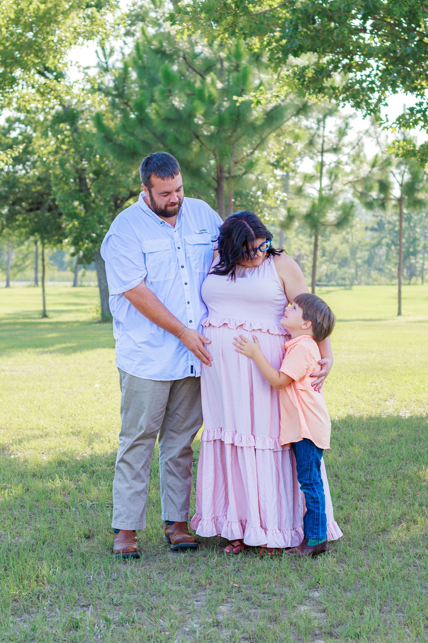 Pregnant woman in pink dress with husband and young son touching her baby bump in a sunny green park.