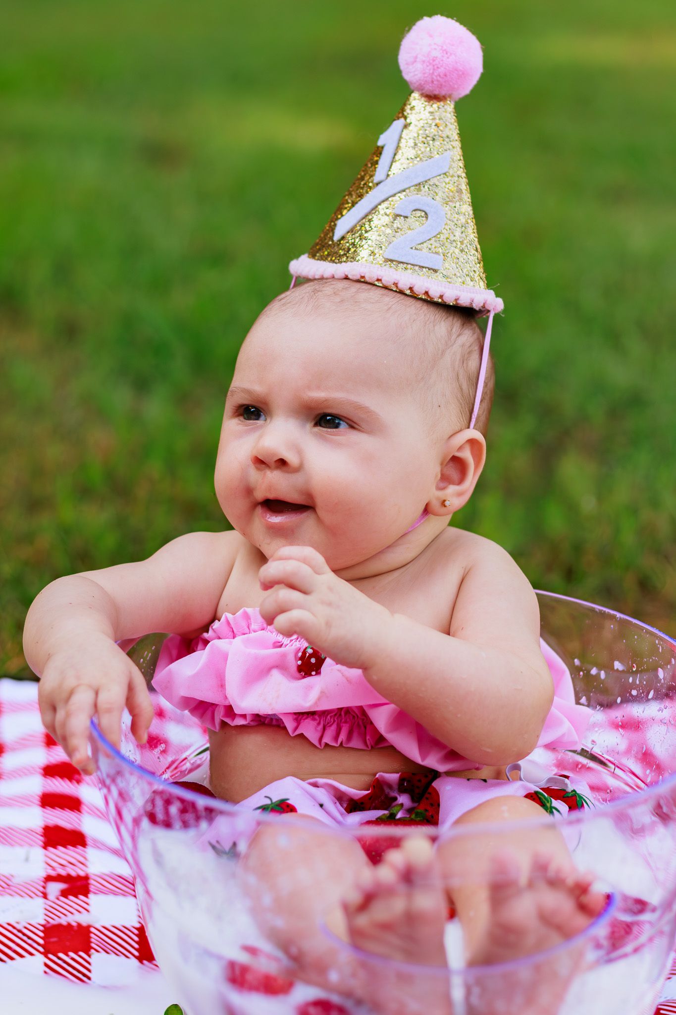 Cute baby girl in pink ruffle outfit wearing gold glitter half birthday party hat sitting outdoors on checkered blanket.