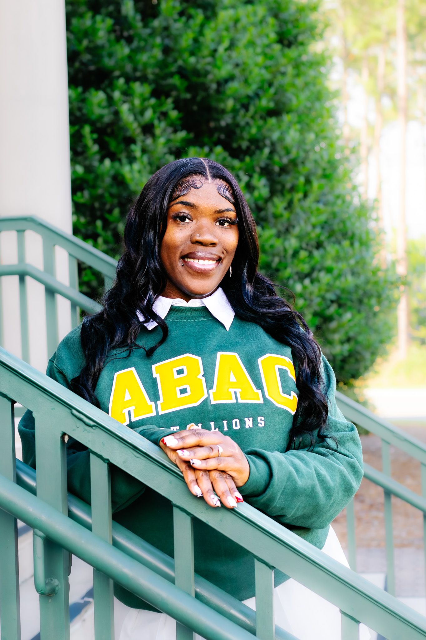 Smiling young woman in green and gold ABAC Stallions sweatshirt leaning on stair railing outdoors.