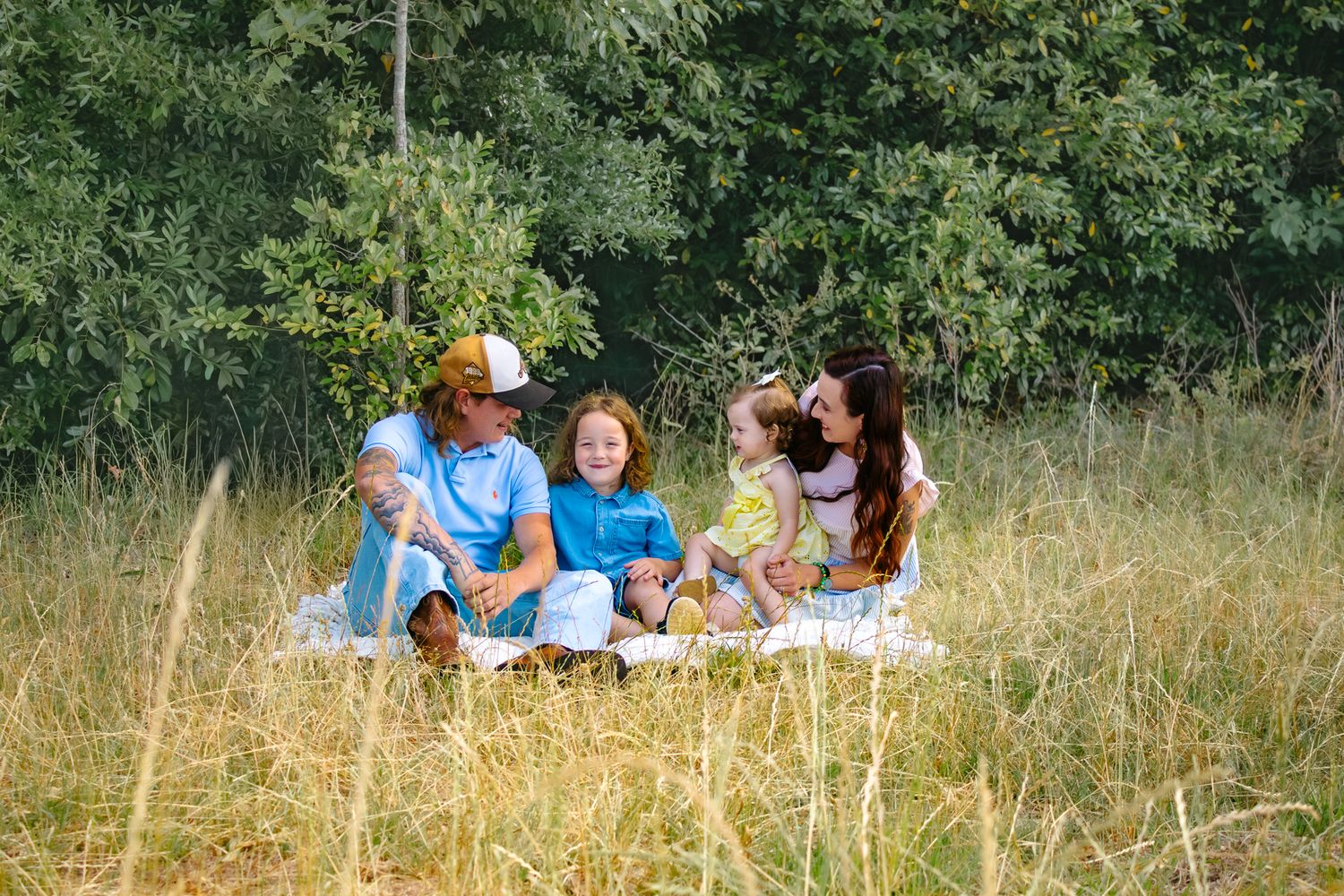 Family of four enjoying a picnic in a golden grass field surrounded by lush green trees on a sunny day.