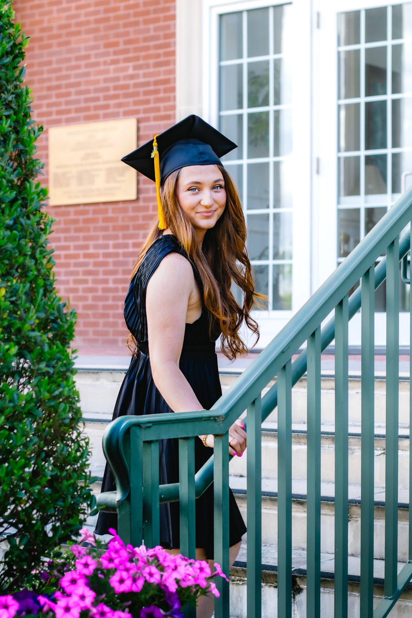 Smiling female graduate in cap and gown poses on staircase with pink flowers and brick building backdrop.