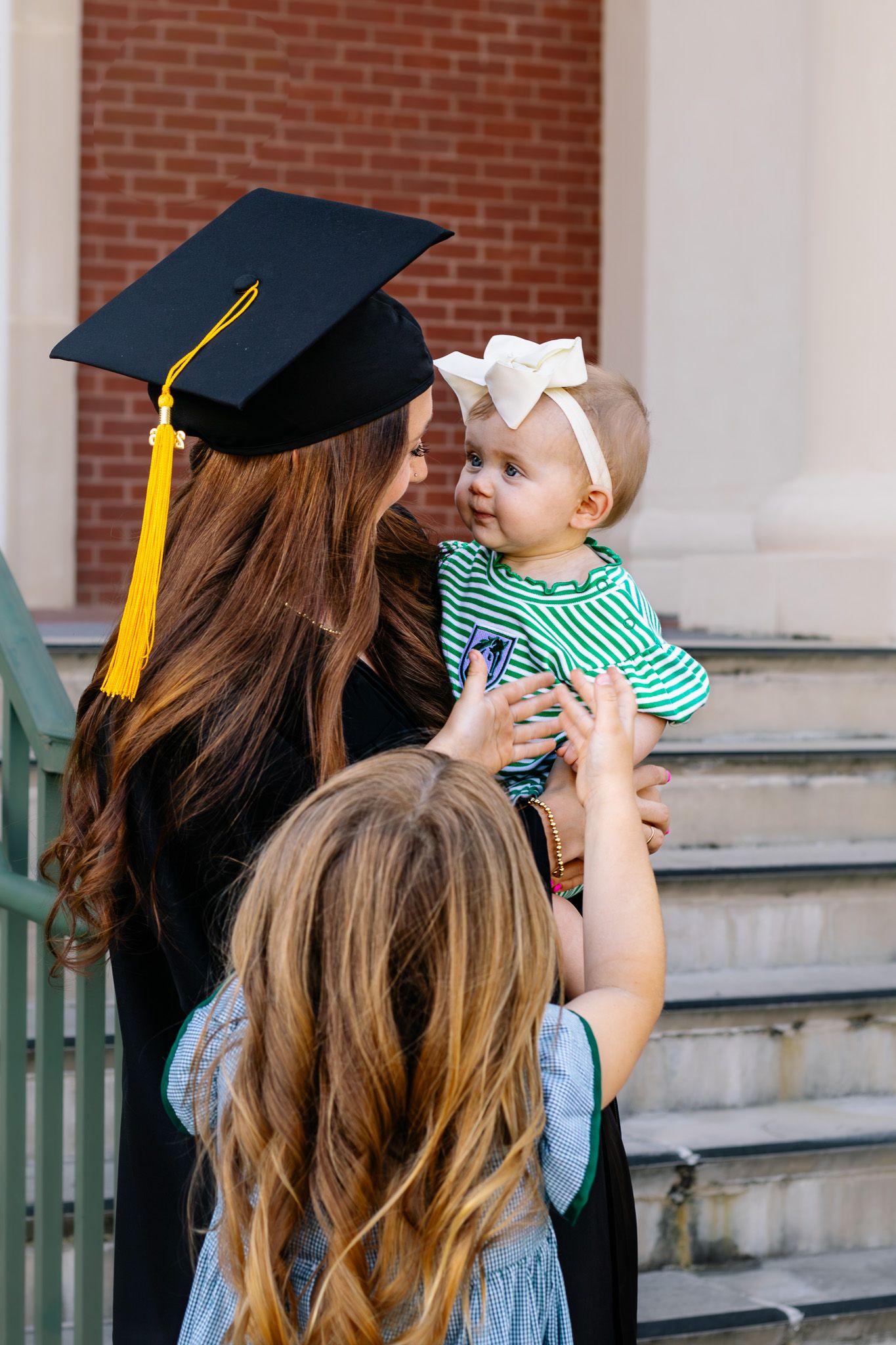Graduate in cap and gown holding baby while child reaches up on steps.