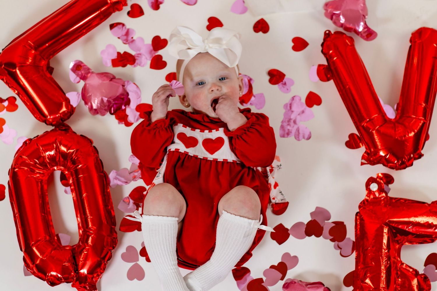 Baby girl in red heart romper lying among LOVE balloons and colorful heart confetti for Valentine's Day.