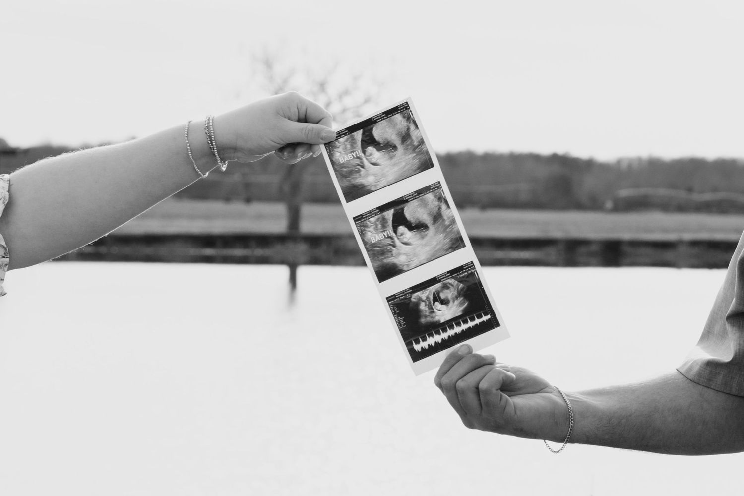 Two hands holding ultrasound photos in black and white outdoor pregnancy announcement.