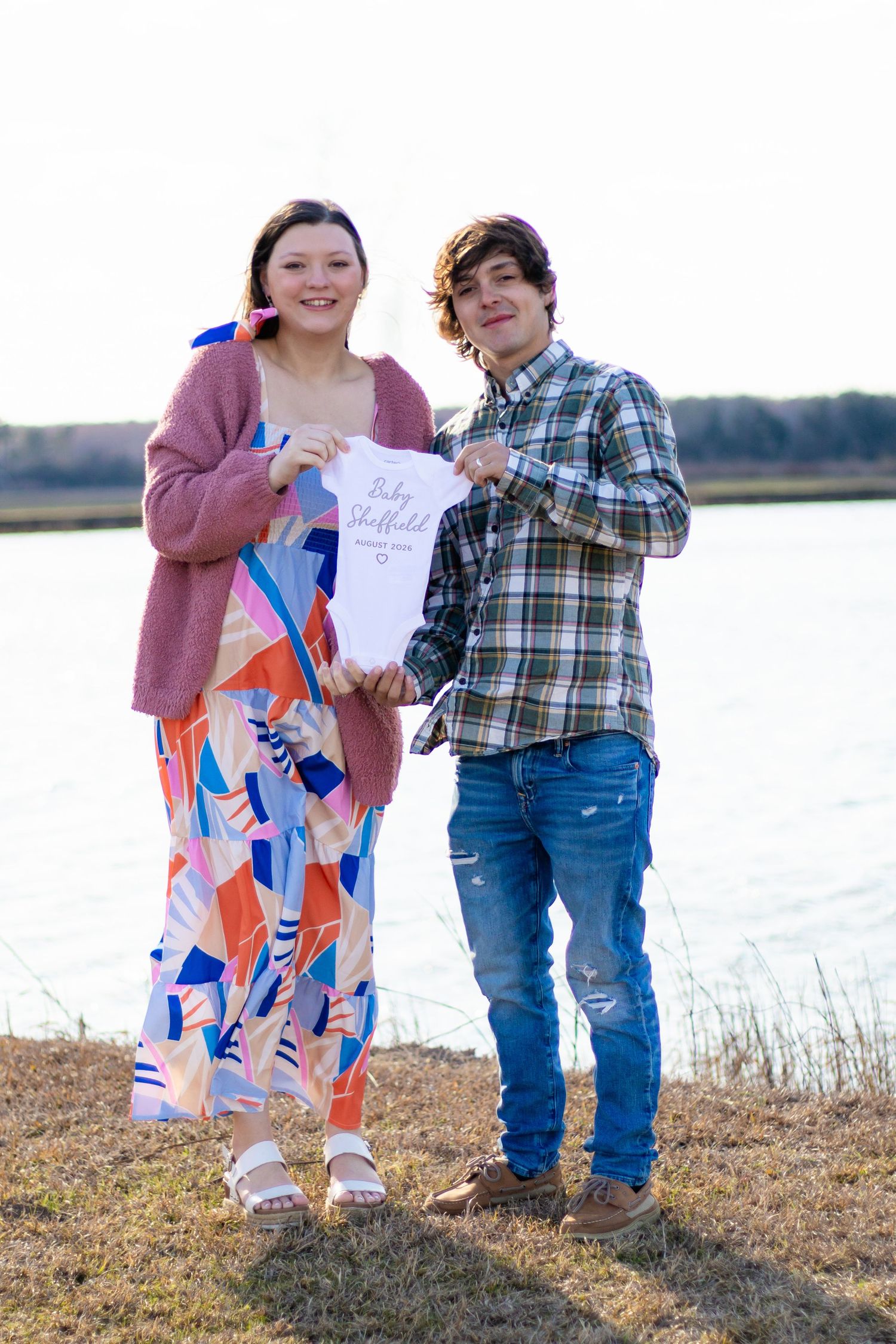 Couple standing by lake holding Baby Sheffield onesie for pregnancy announcement.