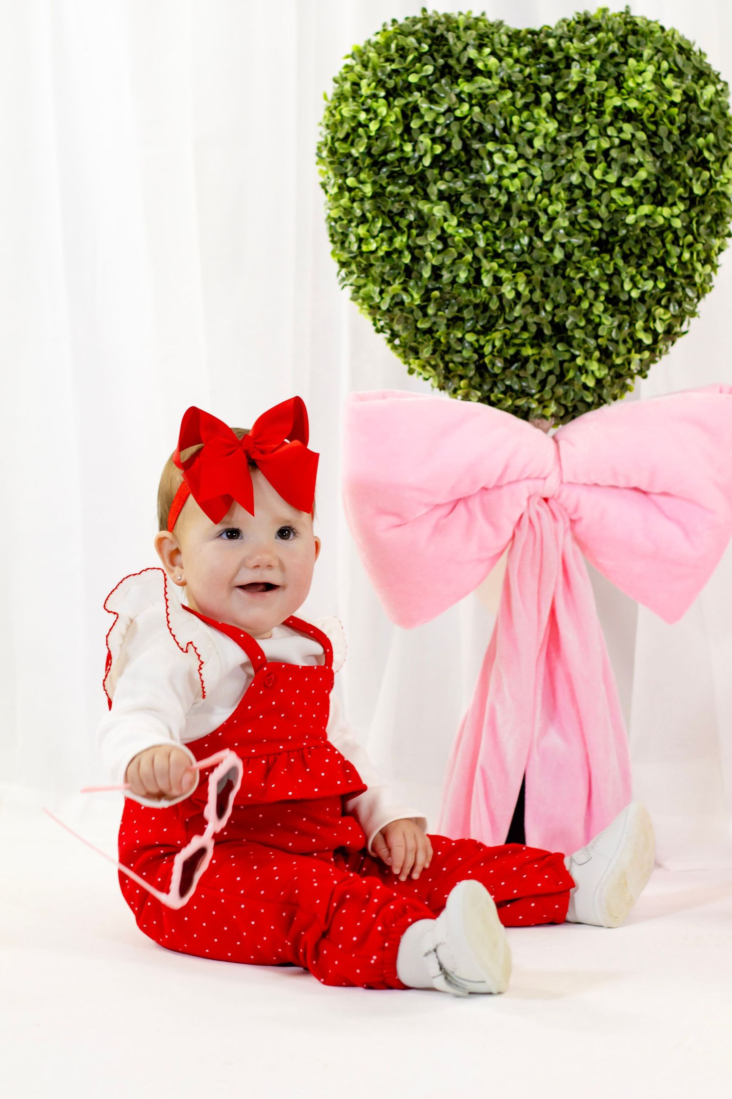 Smiling baby girl in red polka dot romper with bow headband sits beside a heart-shaped topiary.