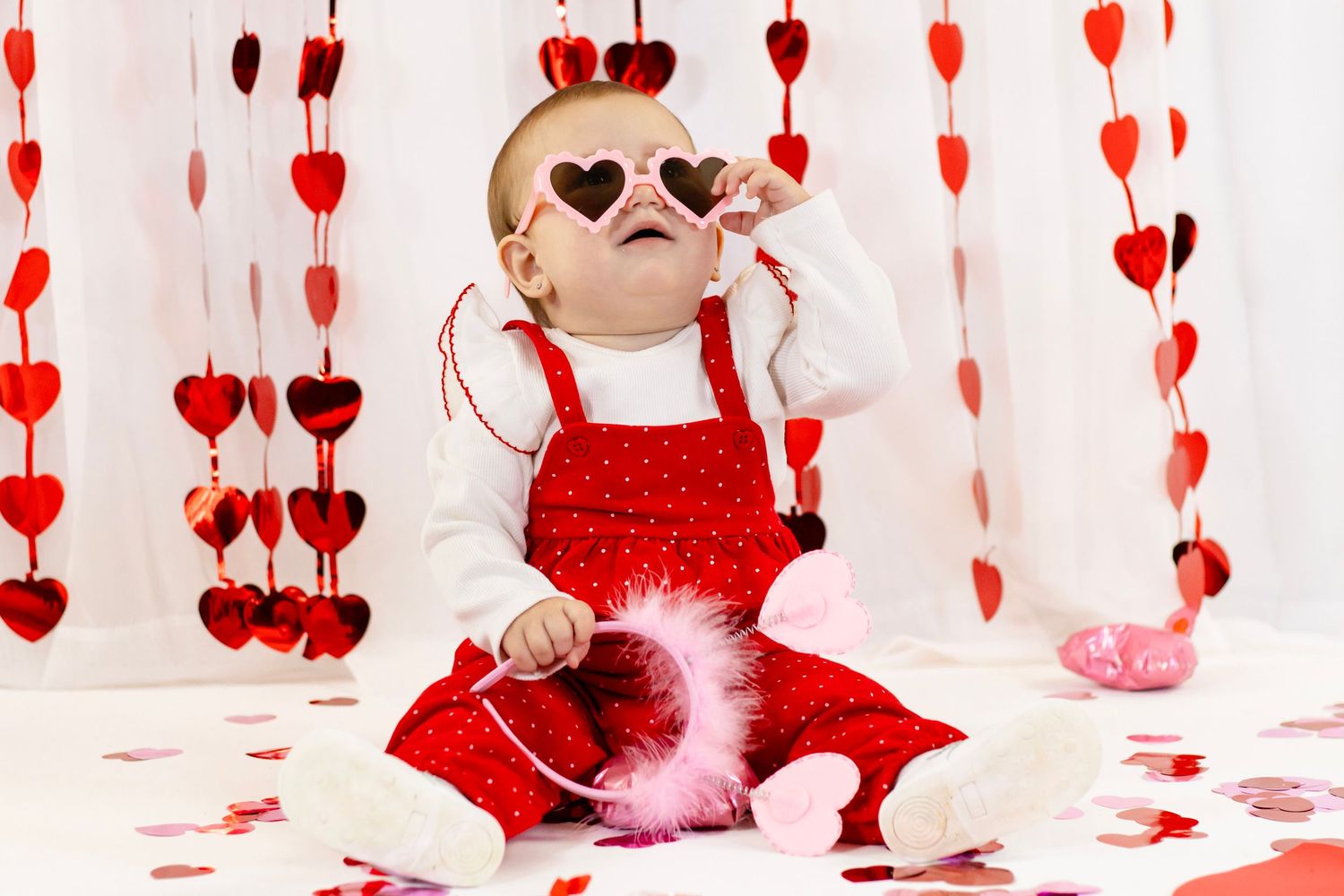 Baby in red polka dot outfit and heart sunglasses plays with pink feather boa among Valentine's Day decorations.