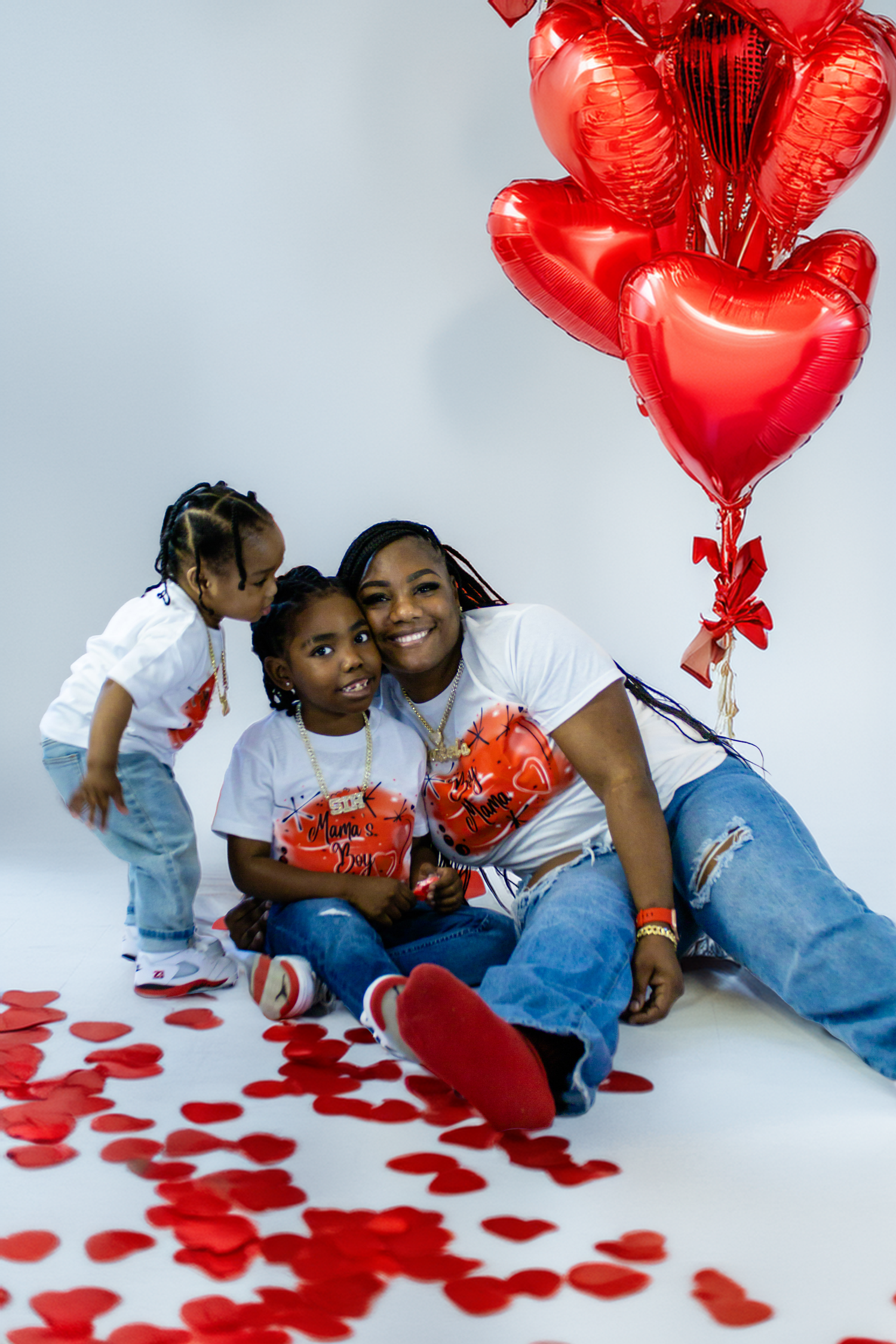 Mom and kids sit on floor surrounded by red heart confetti and heart-shaped balloons for Valentine's Day.