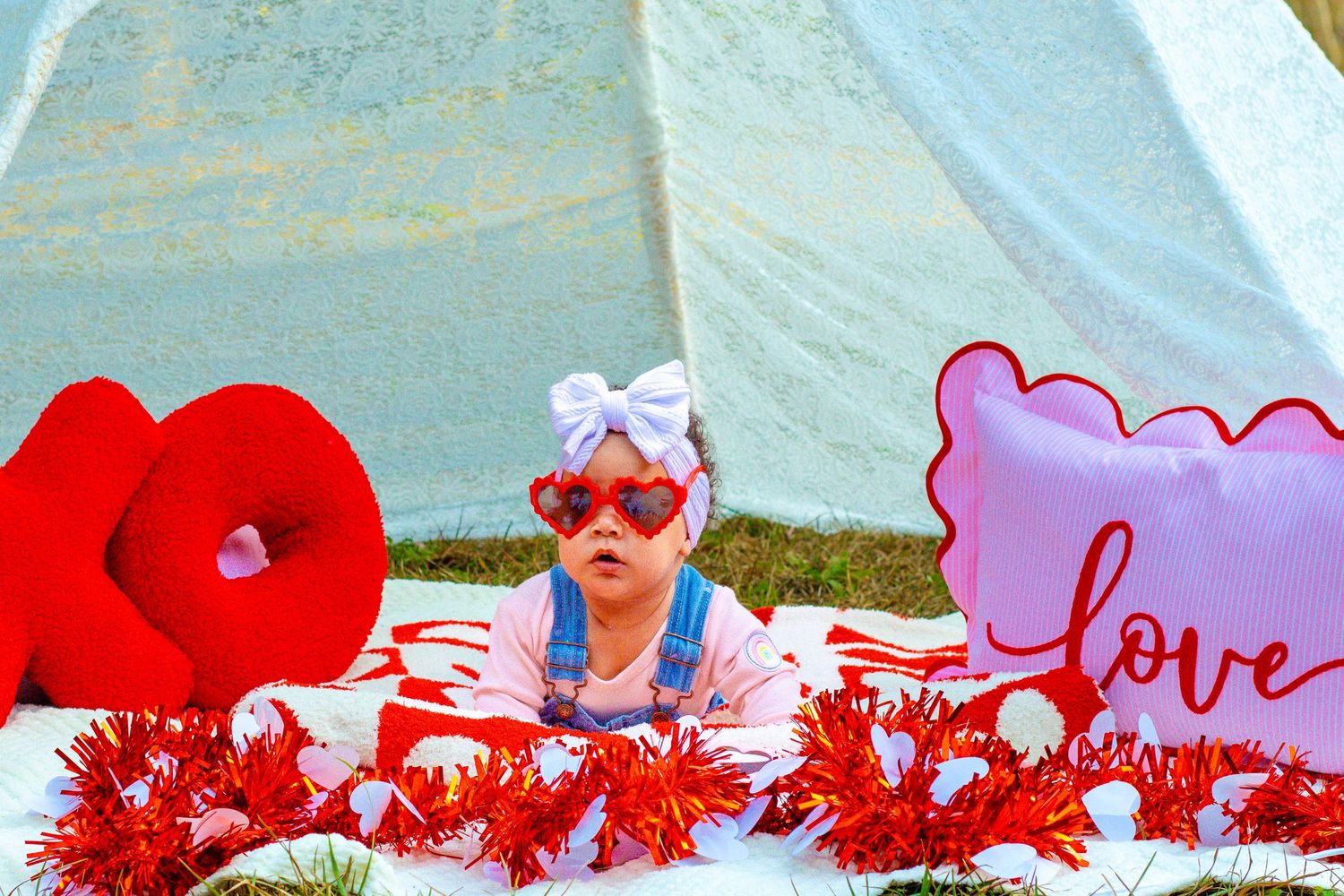 Baby in heart sunglasses and blue outfit peeks over Valentine decorations in front of white teepee outdoors.
