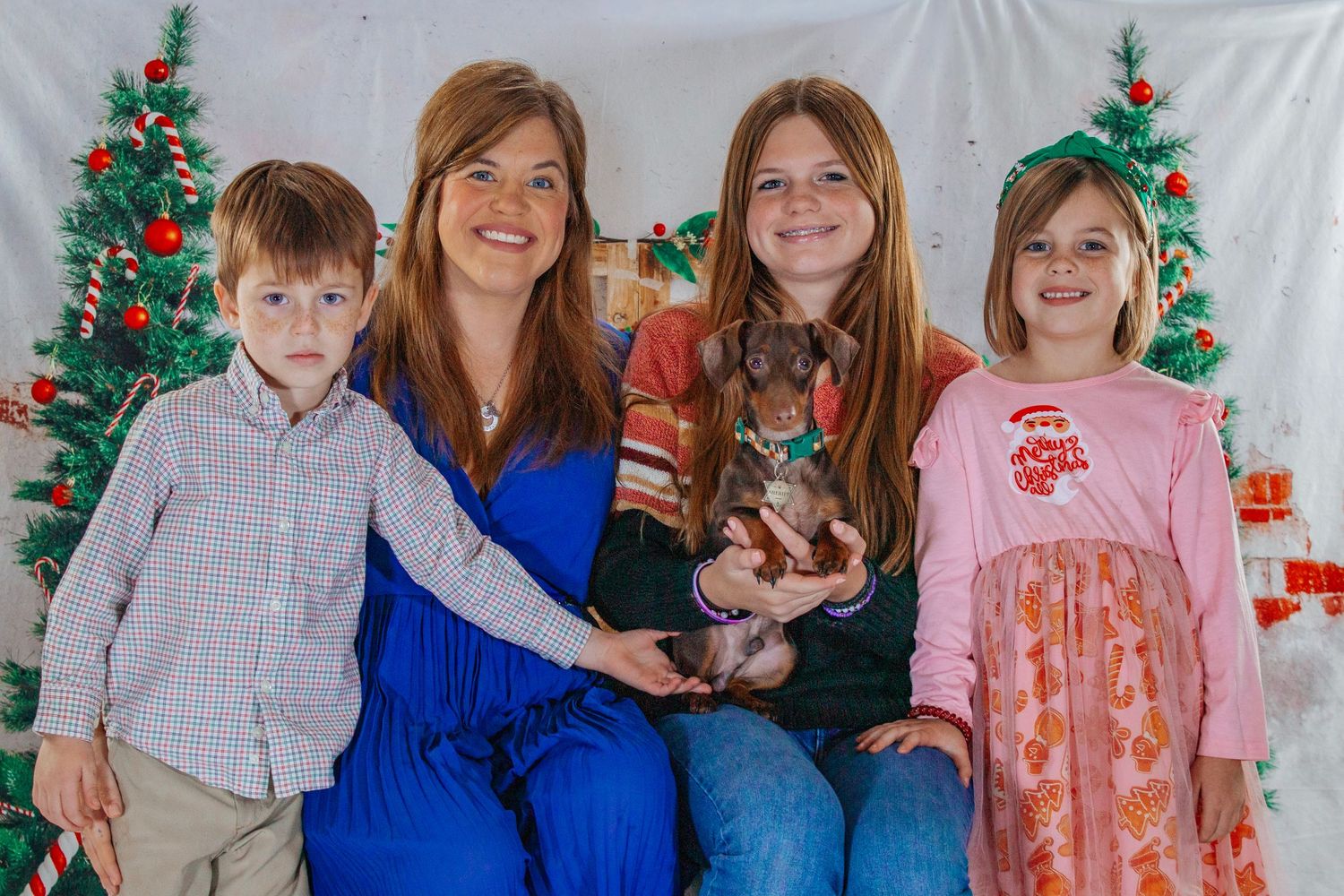 A woman and three children smile together holding a small dachshund in a Christmas-decorated studio setting.