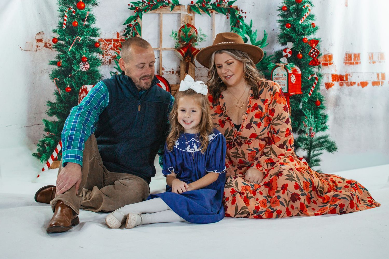 A family of three sits together smiling in front of a festive Christmas tree backdrop indoors.