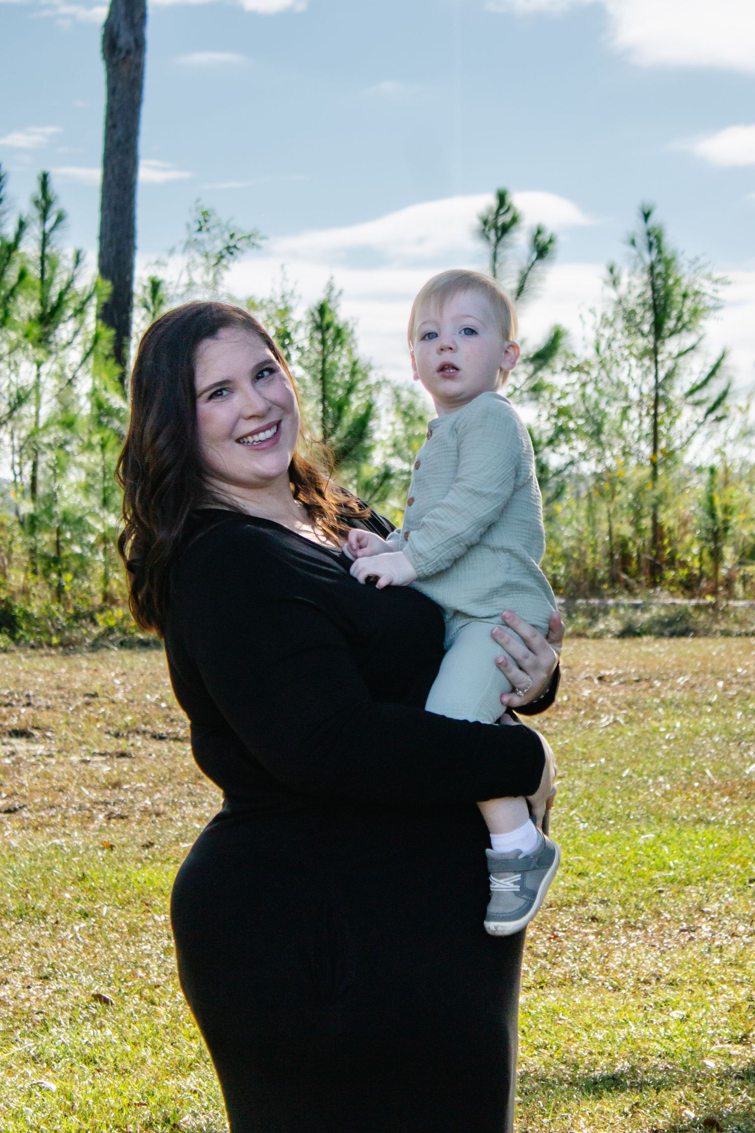 Smiling pregnant woman in black dress holding blonde toddler outdoors in sunny park.