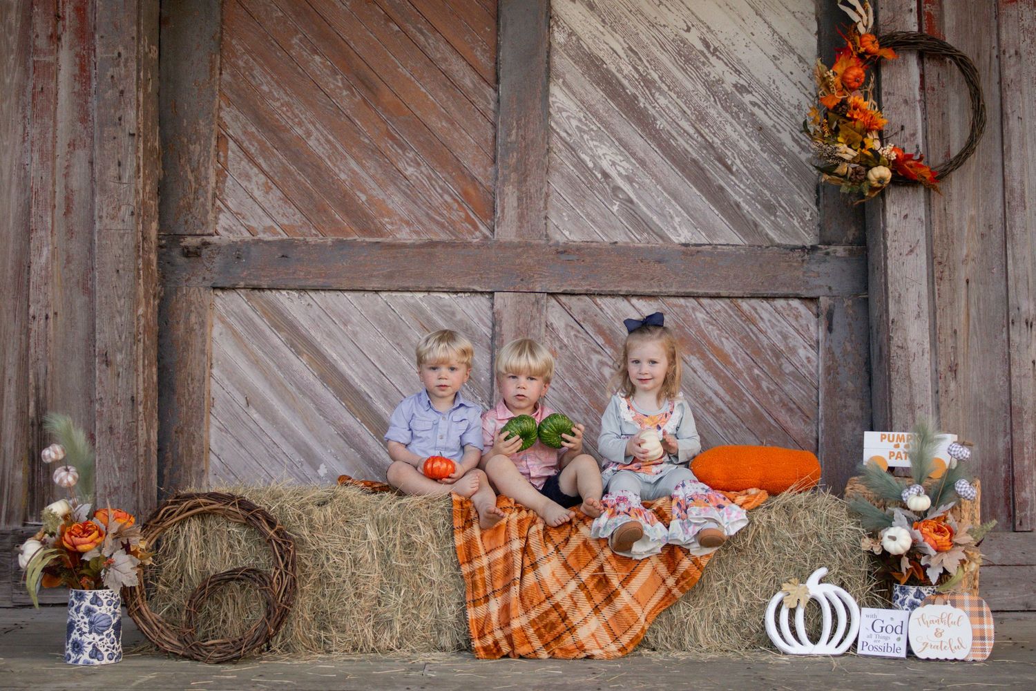 Three young children sit on hay bales with fall pumpkin decorations in front of a rustic barn door.