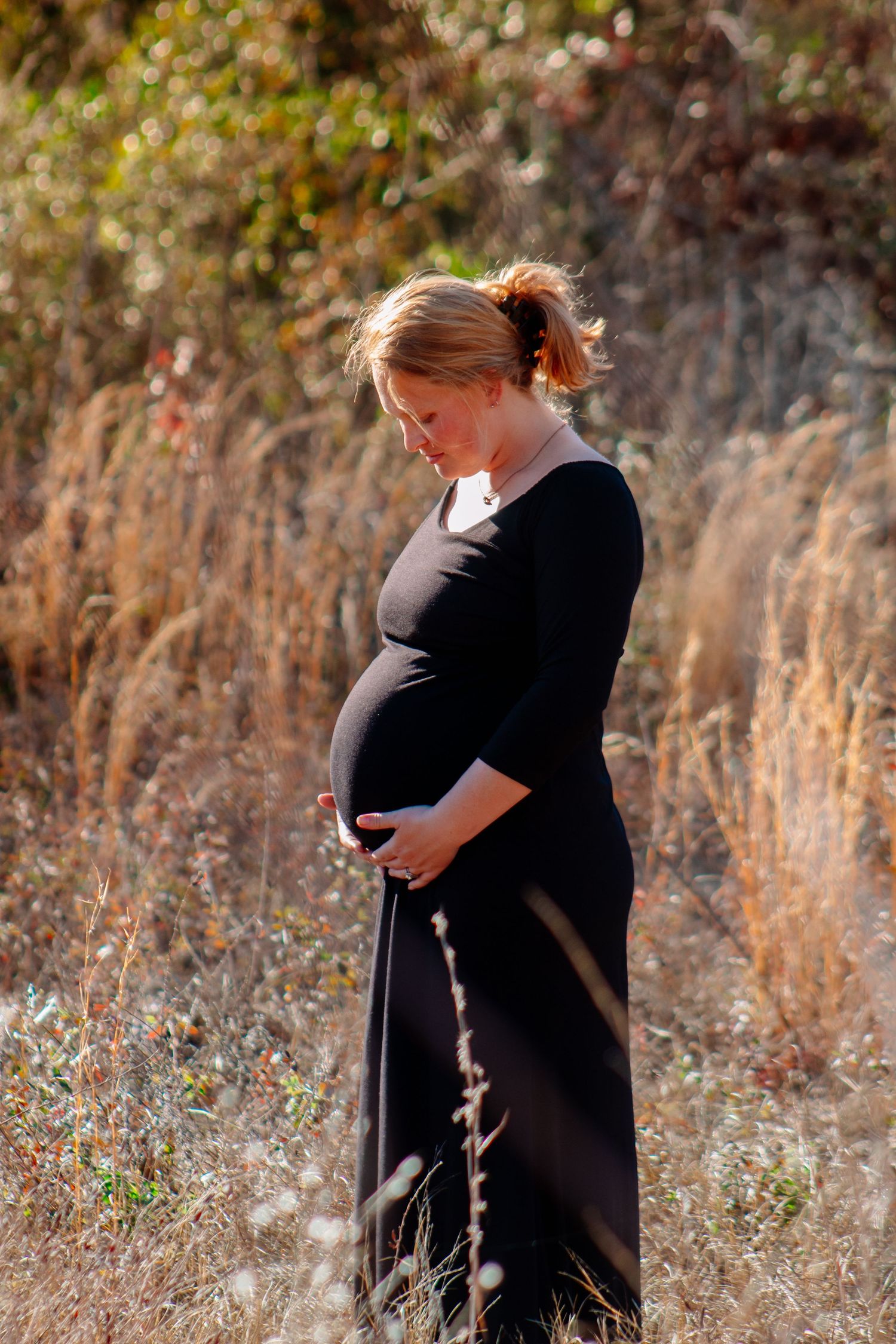 Pregnant woman in black maxi dress cradling baby bump while standing in golden autumn field at sunset.