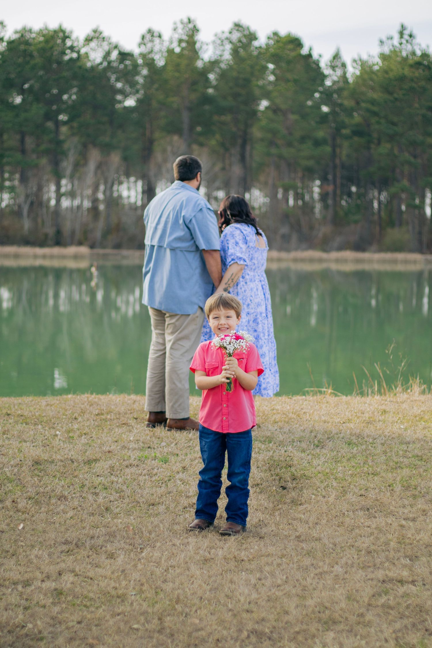 A smiling boy in a red shirt holds flowers near a lake as his parents stand together in the background.