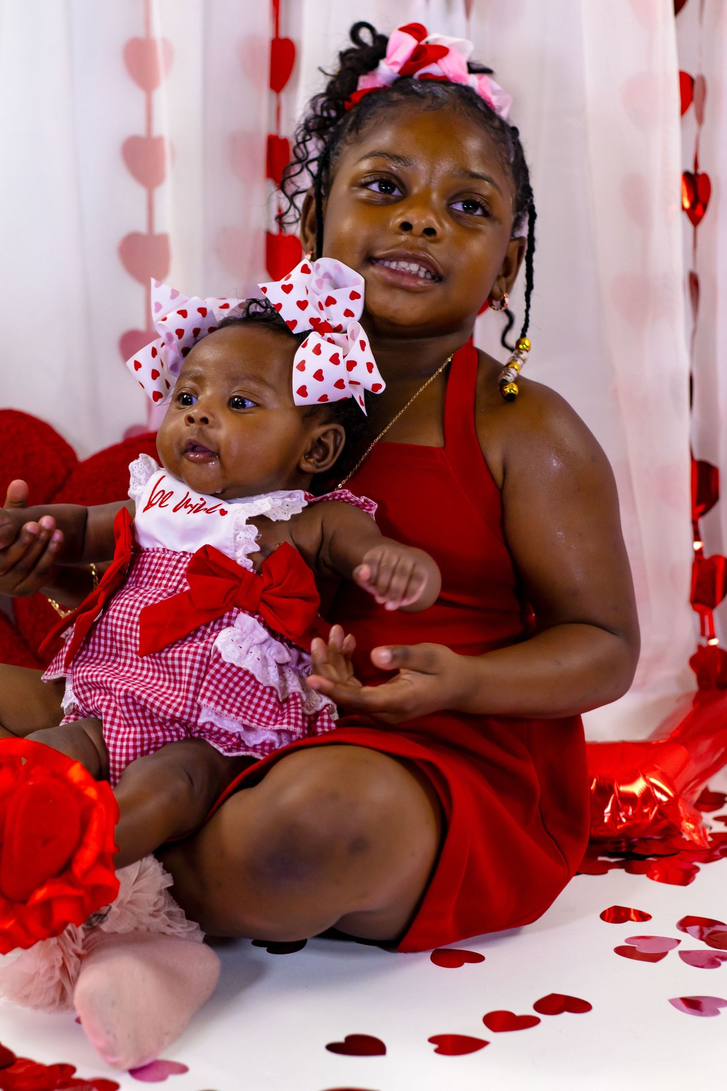 Older sister in red dress holding baby in Valentine's outfit among heart decorations and confetti.