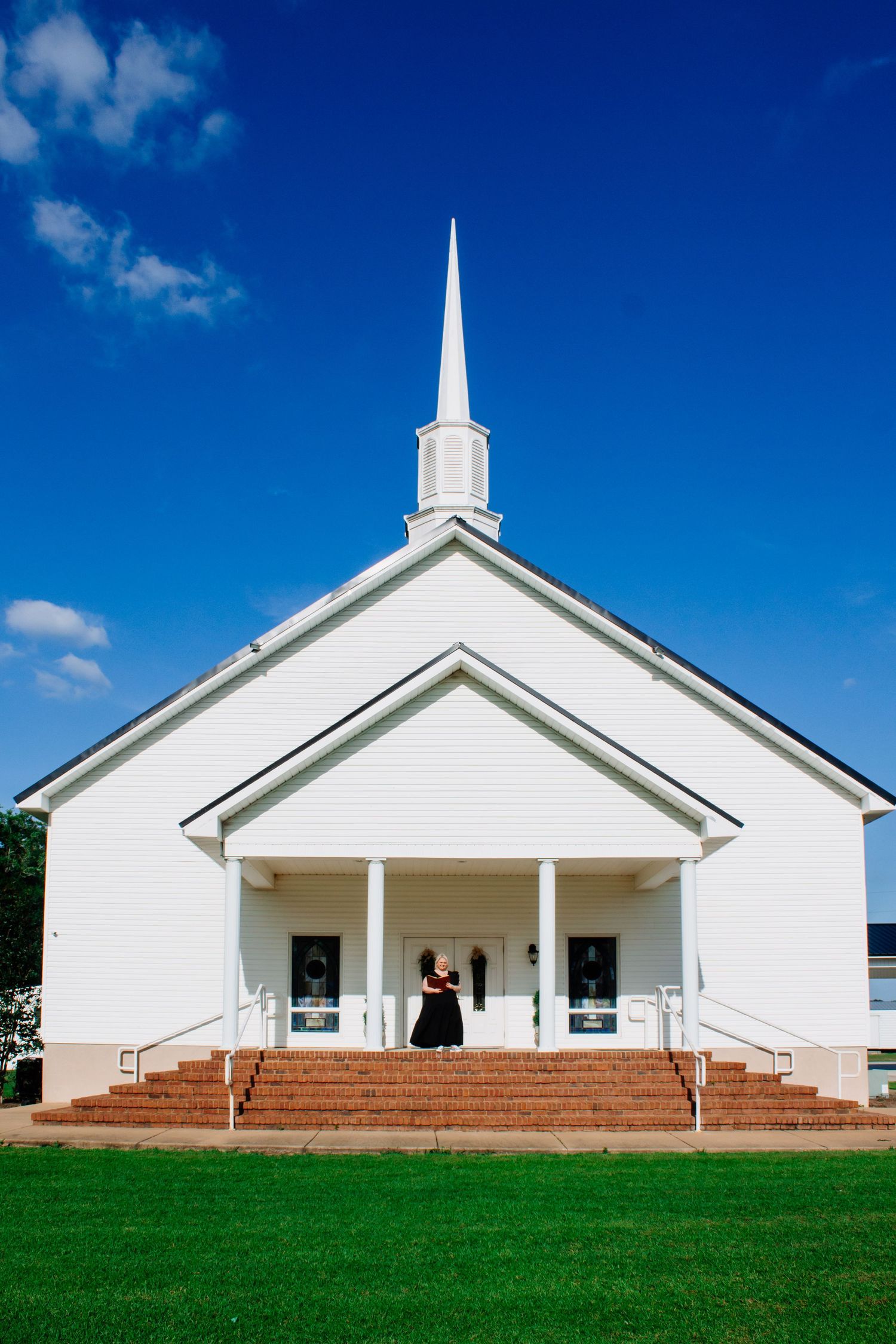 White traditional church with tall steeple against blue sky with woman in black dress on front steps.
