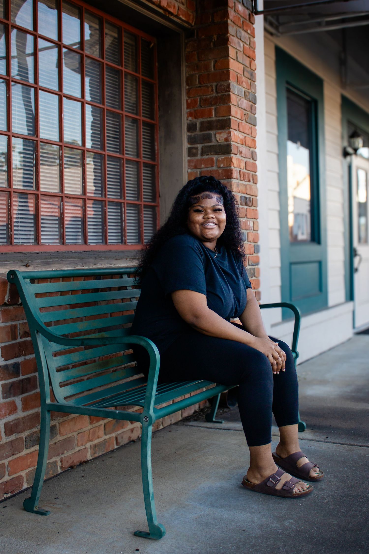 Young Black woman in black outfit smiles while seated on a green bench outside a brick building.