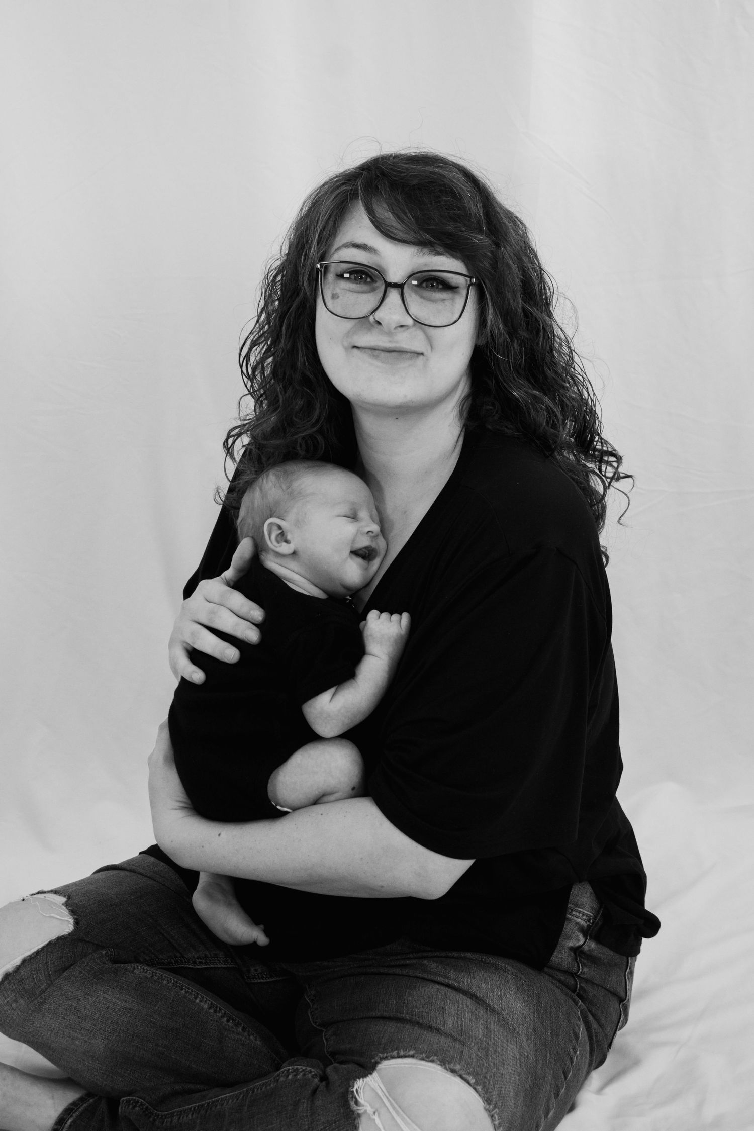 Black and white photo of a curly-haired mother with glasses sitting and cradling her sleeping newborn.