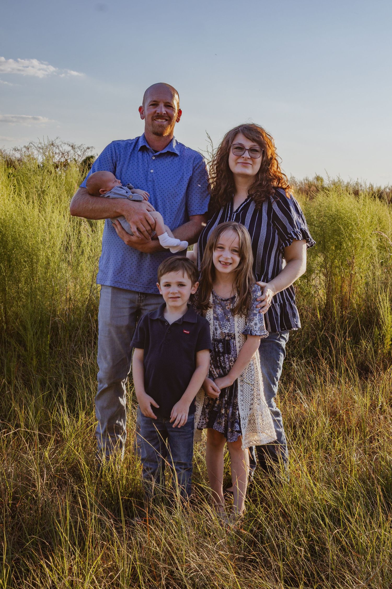 Family of five with newborn posing in golden hour sunlight among tall green grass in field.