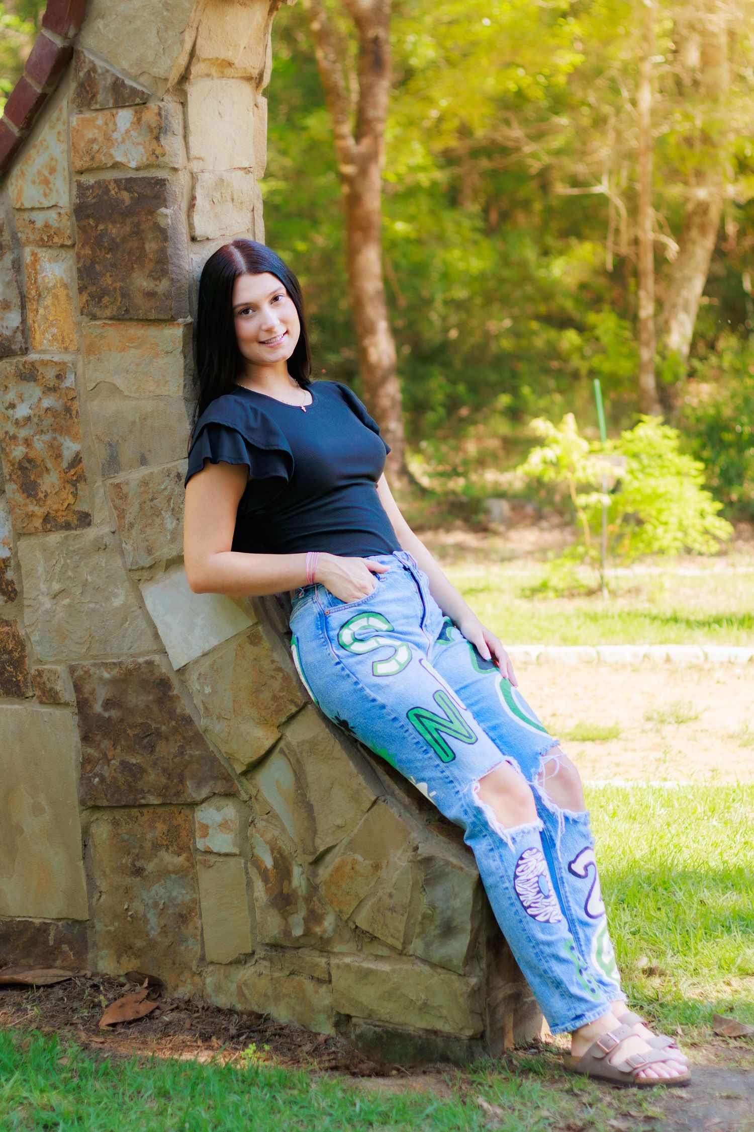 Young woman in decorated jeans and black top leaning against stone wall in park.