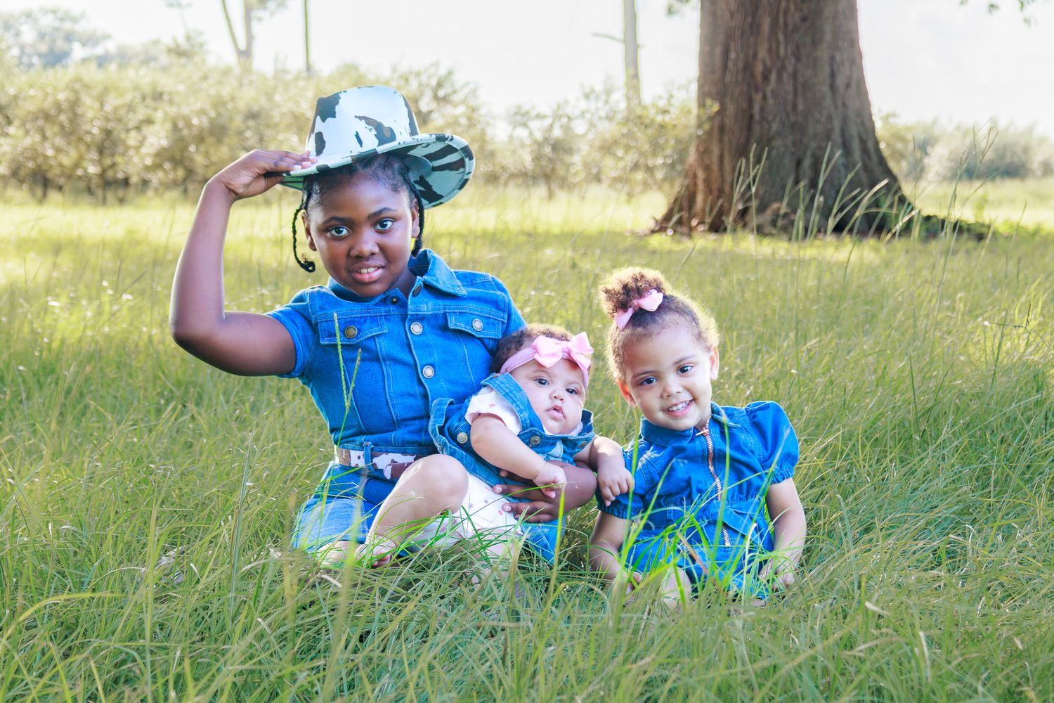 Three young children in matching blue outfits sitting in green grass near a tree outdoors.