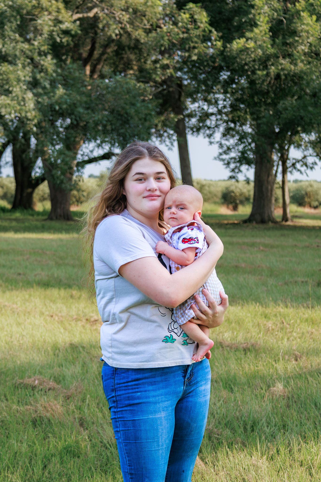 Young woman holding a newborn baby outdoors in a green field with trees in the background.