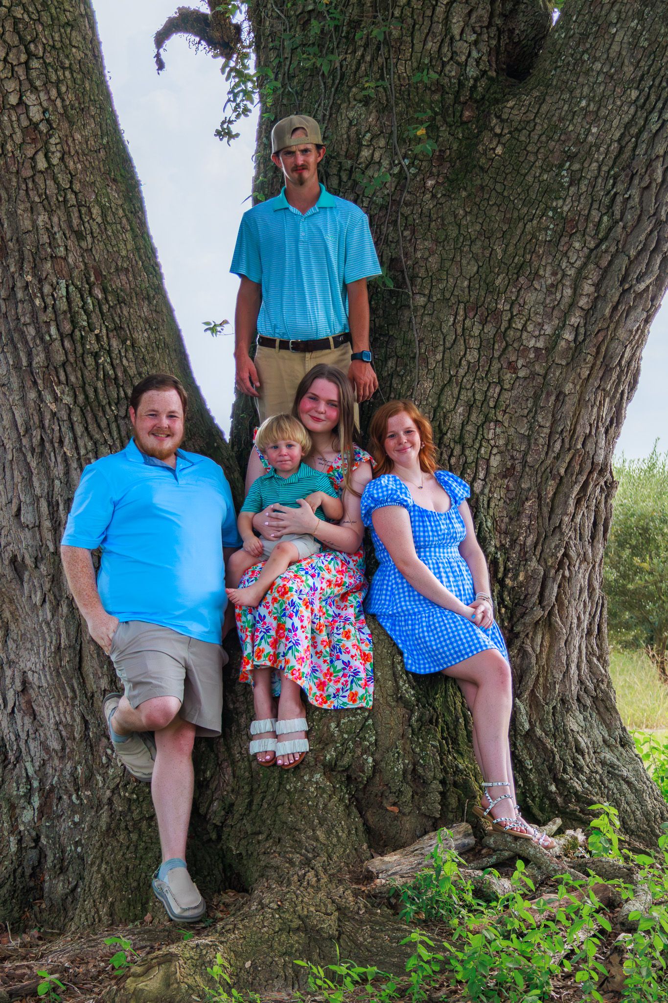 Family of five posing together around a large tree trunk outdoors on a summer day.