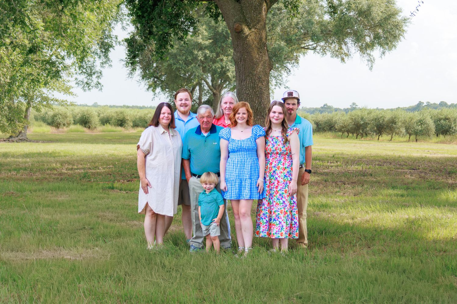 Family of seven posing outdoors under a large tree in a green field, dressed in colorful summer attire.