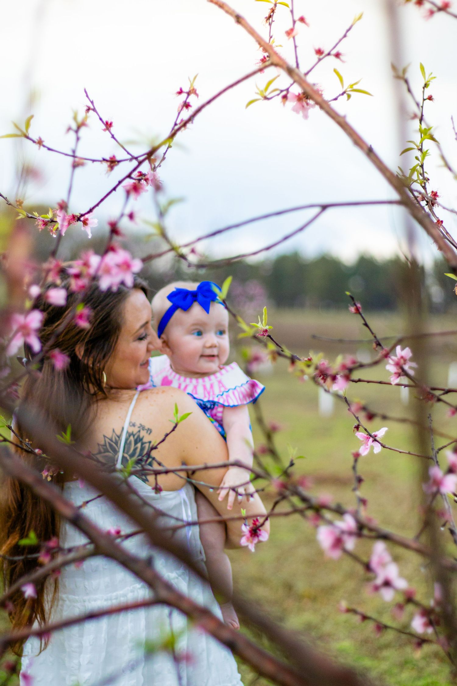Mother holds smiling baby girl with blue bow headband among pink flowering tree branches in spring orchard.